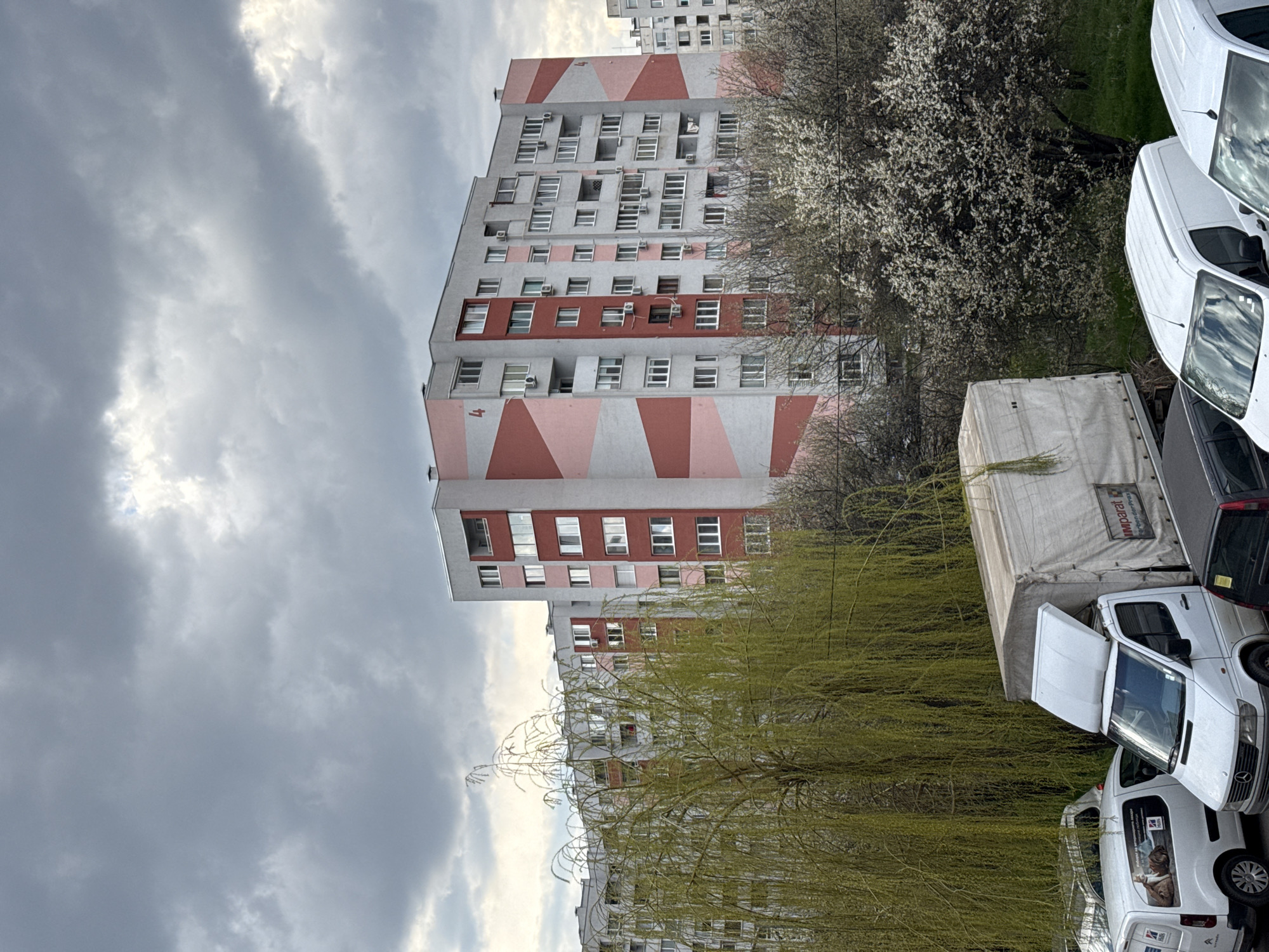 This image depicts a residential scene in Sofia, Bulgaria, under a heavily overcast, dark grey sky. The dominant features are several multi-story apartment buildings. The most prominent building, centrally located, is a concrete block structure with at least eight visible stories, painted in a distinctive pattern of light grey, light pink, and reddish-pink diagonal stripes and solid panels. The number "4" is painted on the building's facade in two locations. Numerous windows and multiple external air conditioning units are visible on this and surrounding buildings. Laundry is visible hanging on some balconies. In the foreground, a large weeping willow tree with bright green, newly unfurled leaves occupies the left side, partially obscuring the buildings. To its right, a tree covered in white blossoms is visible. Below the trees, several vehicles are parked on what appears to be a grassy or unpaved area. A large white cargo truck with a grey canvas cover, displaying "IMPARAT" and "V.M. Derzhavin" in Cyrillic, is centrally positioned with its hood partially open. To its left, a white Citroën van is parked, featuring "Hills" branding and the Bulgarian text "За вашето ветеринарно здраве" (For your veterinary health) alongside images of a dog and cat. Partially visible to the right of the large truck are the fronts of at least two other white vans and a dark-colored car. No people are visible in the scene.