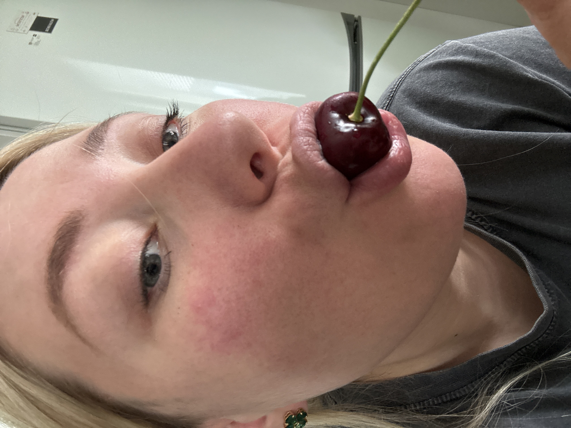 In an indoor setting, a close-up, eye-level shot captures a person's face from the chin up, with a cherry held between their lips. The subject has light-colored hair and is wearing a dark grey t-shirt. The background is out of focus, with a white surface on the right and a hint of blonde hair on the left. The lighting appears to be natural, coming from the front. The image has a shallow depth of field, focusing on the subject's mouth and the cherry.