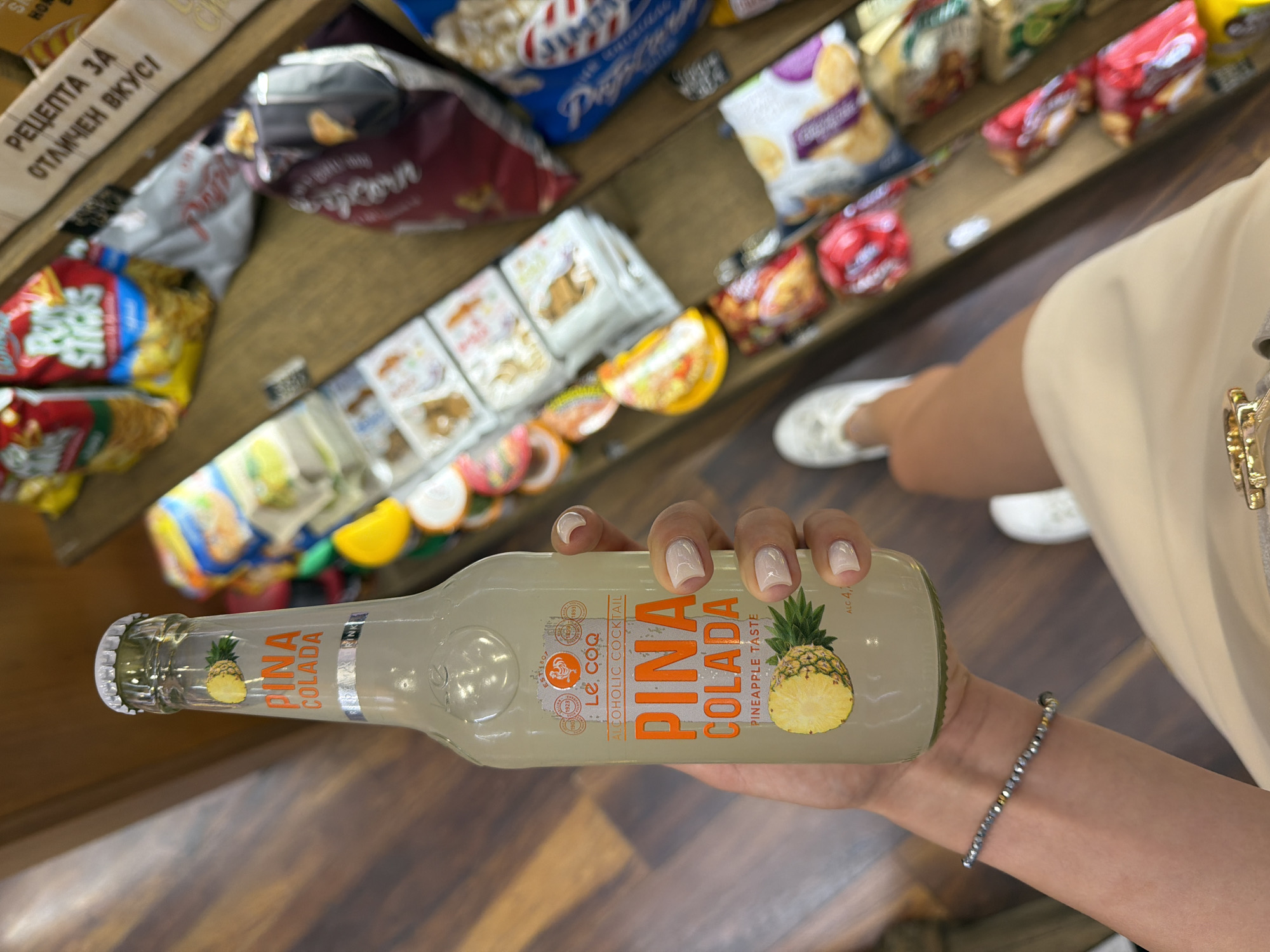A person's arm and hand are holding a bottle of "Pina Colada" alcoholic cocktail. The bottle is clear glass and contains a pale yellow liquid. The label on the bottle features an illustration of a pineapple and the words "PINA COLADA" in orange lettering, along with "LE. COQ ALCOHOLIC COCKTAIL" and "PINEAPPLE TASTE". The person's fingernails are painted a light, neutral color. The background consists of shelves stocked with various packaged snacks, including bags of popcorn and other chips. The scene appears to be inside a store or market. There is also a beaded bracelet on the person's wrist. The floor is wooden. There are no visible landmarks or distinguishing features that would allow for precise location identification within Sofia, Bulgaria.