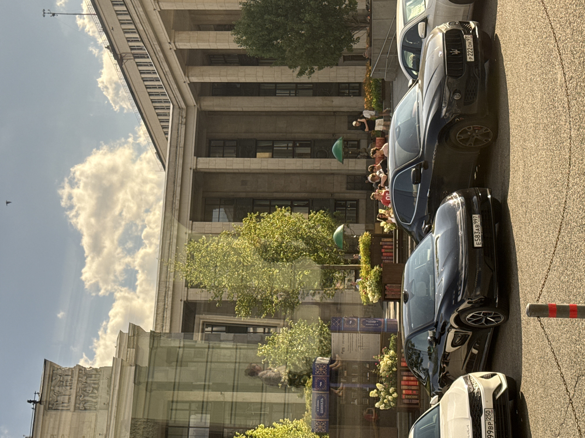 In Moscow, Russia, a daytime, eye-level shot captures a street scene in front of a grand, classically-styled building with tall columns and numerous windows. In the foreground, several cars are parked, including a white car on the left with license plate "99BP 250", a black sedan with license plate "E583AB 997", and a black SUV with license plate "P 222 OH 97". The SUV is a Maserati Levante. A white car is partially visible on the right. In the middle ground, a group of people are gathered, some standing and some sitting, near planters filled with flowers and decorative books. A green umbrella is visible behind some of the people. The background features the imposing architecture of the building, with visible sculptures and detailing on its facade. The sky above is partly cloudy with bright sunlight. The reflection of trees and people is visible on the glass facade of a building to the left.