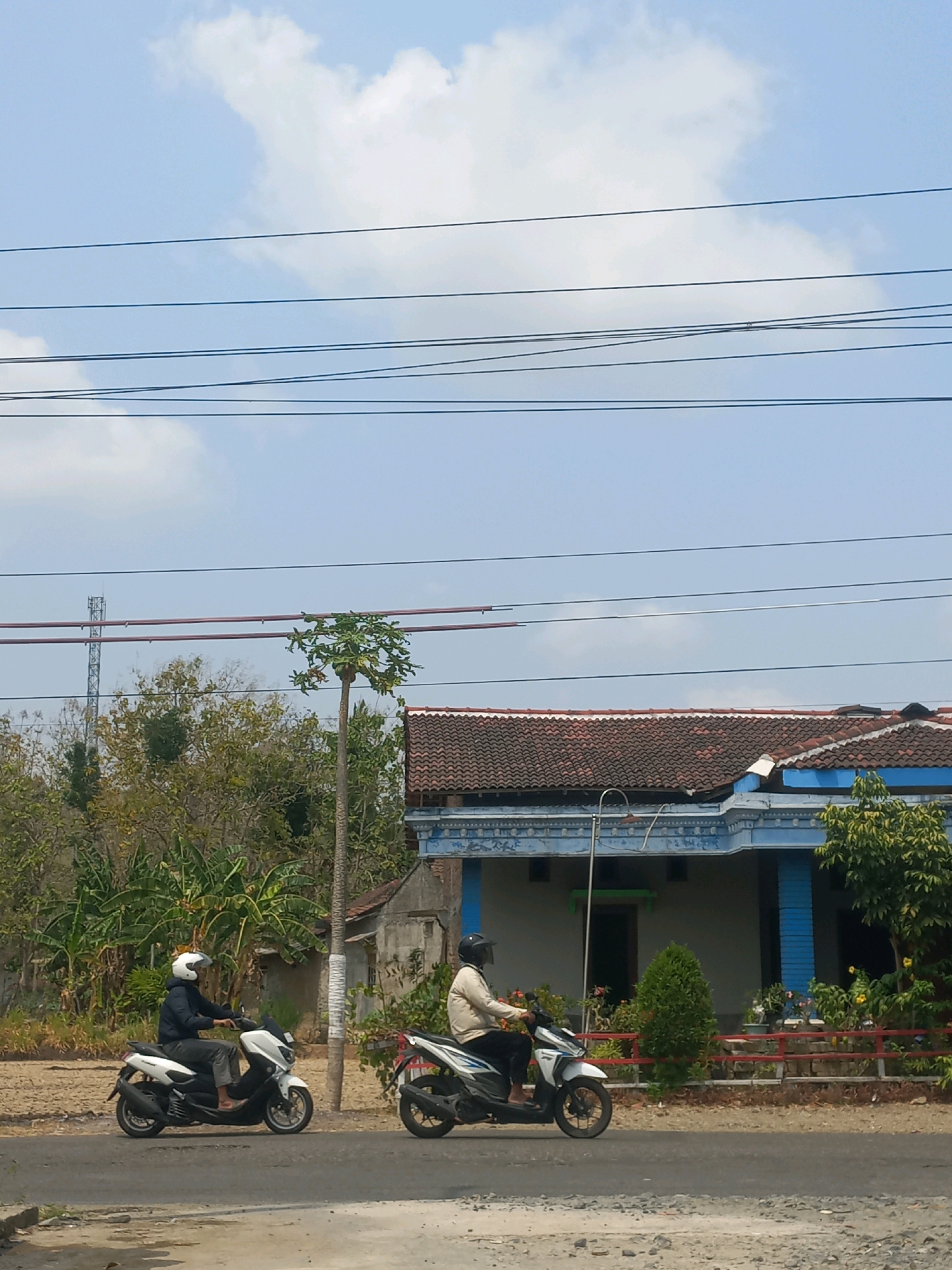 Two individuals are riding scooters on a paved road in Baturetno, Indonesia. The scooter on the left is white with a rider wearing a dark blue jacket and a white helmet. The scooter on the right is white and blue with a rider wearing a light-colored jacket and a black helmet. A house with a red tiled roof and blue trim is visible behind the road. Trees and a tall pole with antennas are also present in the background. Overhead, several power lines stretch across a partly cloudy blue sky.