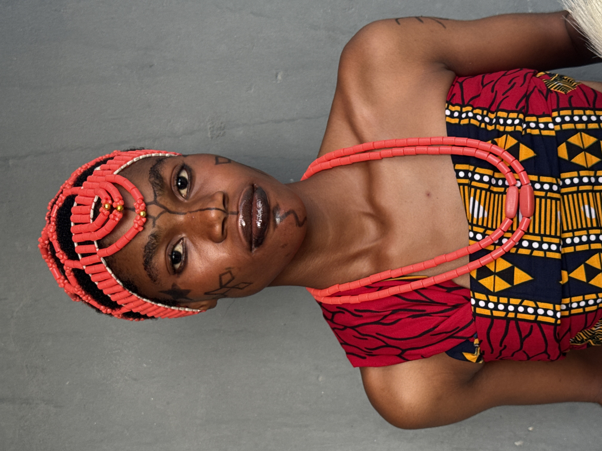 A woman is depicted, possibly in a cultural or ceremonial context in Badagry, Nigeria. She is wearing a headdress composed of rows of coral beads with gold accents, and a matching coral bead necklace. Her face is adorned with markings or tattoos. She wears a red, black, and yellow patterned garment. The background appears to be a plain, gray surface.