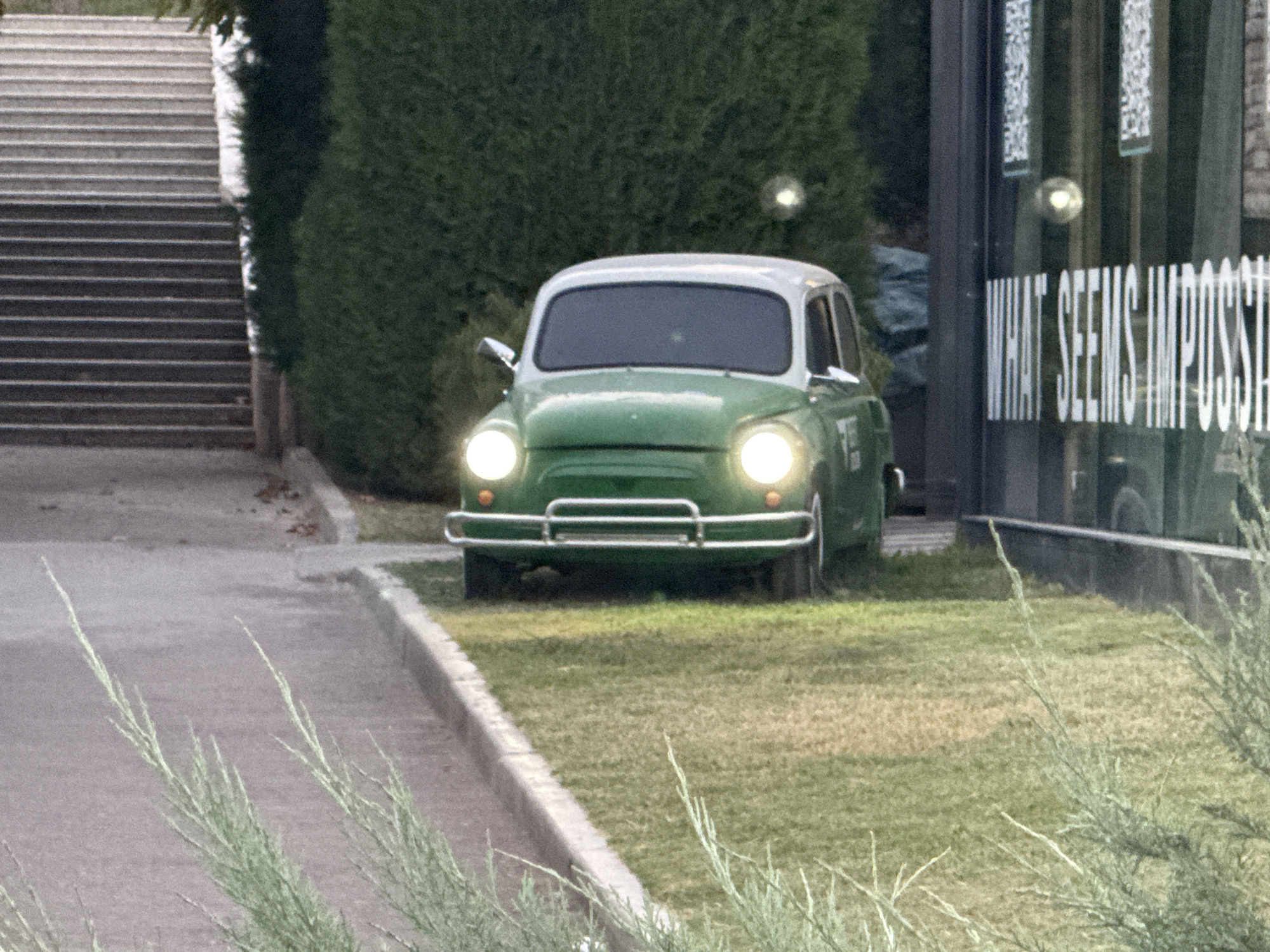 The image shows a vintage green and white car parked on a grassy area next to a building with large windows. The car has its headlights on and a chrome bumper. The building has text on the window that reads "WHAT SEEMS IMPOSSIBLE". To the left of the car are a set of stone stairs and a dense green hedge. In the foreground, there are some dry grass and thin green branches. The overall scene suggests an outdoor setting in an urban environment, possibly a cafe or a business in Tashkent, Uzbekistan, given the presence of the green hedges and building style which are common in the city.