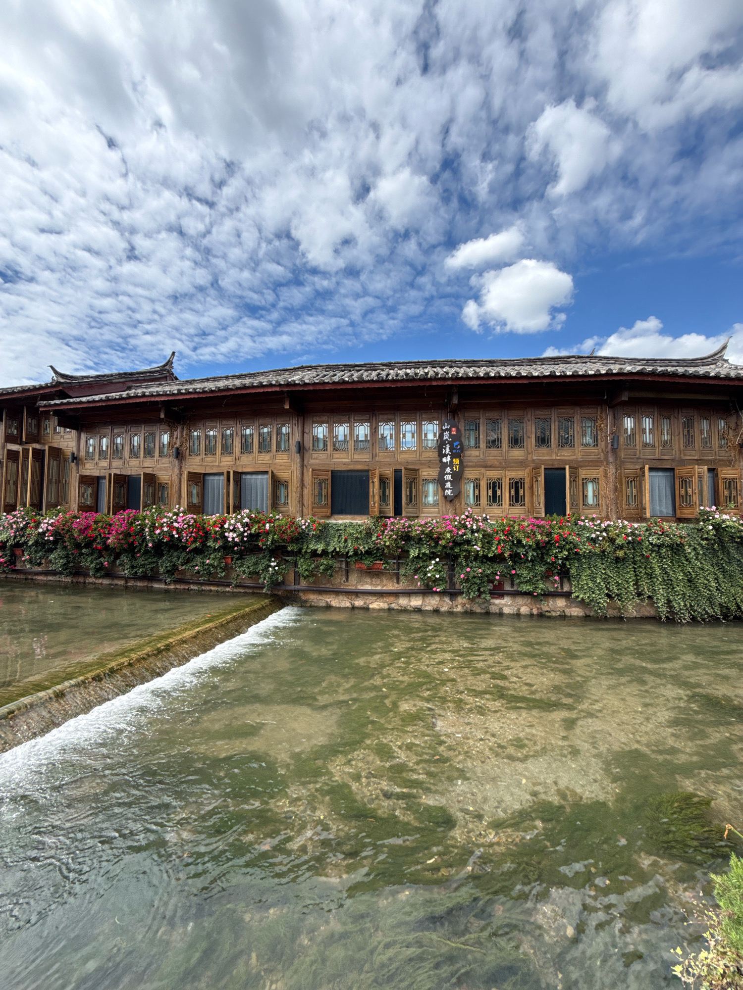 This is a wide, low-angle shot of a historic, wooden building with a curved facade, situated alongside a body of water. The building is two stories tall, with a dark, tiled, curved roofline. The facade is made of light-colored wood with numerous windows on both floors, many of which have wooden shutters that are partially open. The windows on the second floor are all identical, featuring intricate wooden lattice work. Along the first floor, a vibrant display of flowers in shades of pink, red, and white spills over from planters, with lush green ivy growing below them, cascading down towards the water.
The body of water in the foreground is shallow and appears to be a river or canal. A low concrete barrier creates a small waterfall or spillway on the left side of the image, with clear water flowing over it. The water's surface is slightly rippled, and the bottom is visible in places, showing sand, stones, and green aquatic plants.
Above the building, a bright blue sky is dotted with fluffy white clouds. A dark, vertical wooden sign with Chinese characters is visible on the building's facade, to the left of center. The scene suggests a well-preserved, traditional architectural style, likely in a scenic setting. The building appears to be a shop, restaurant, or guesthouse, given its elaborate facade and floral decorations. This location is in the city of Gucheng District, China.
