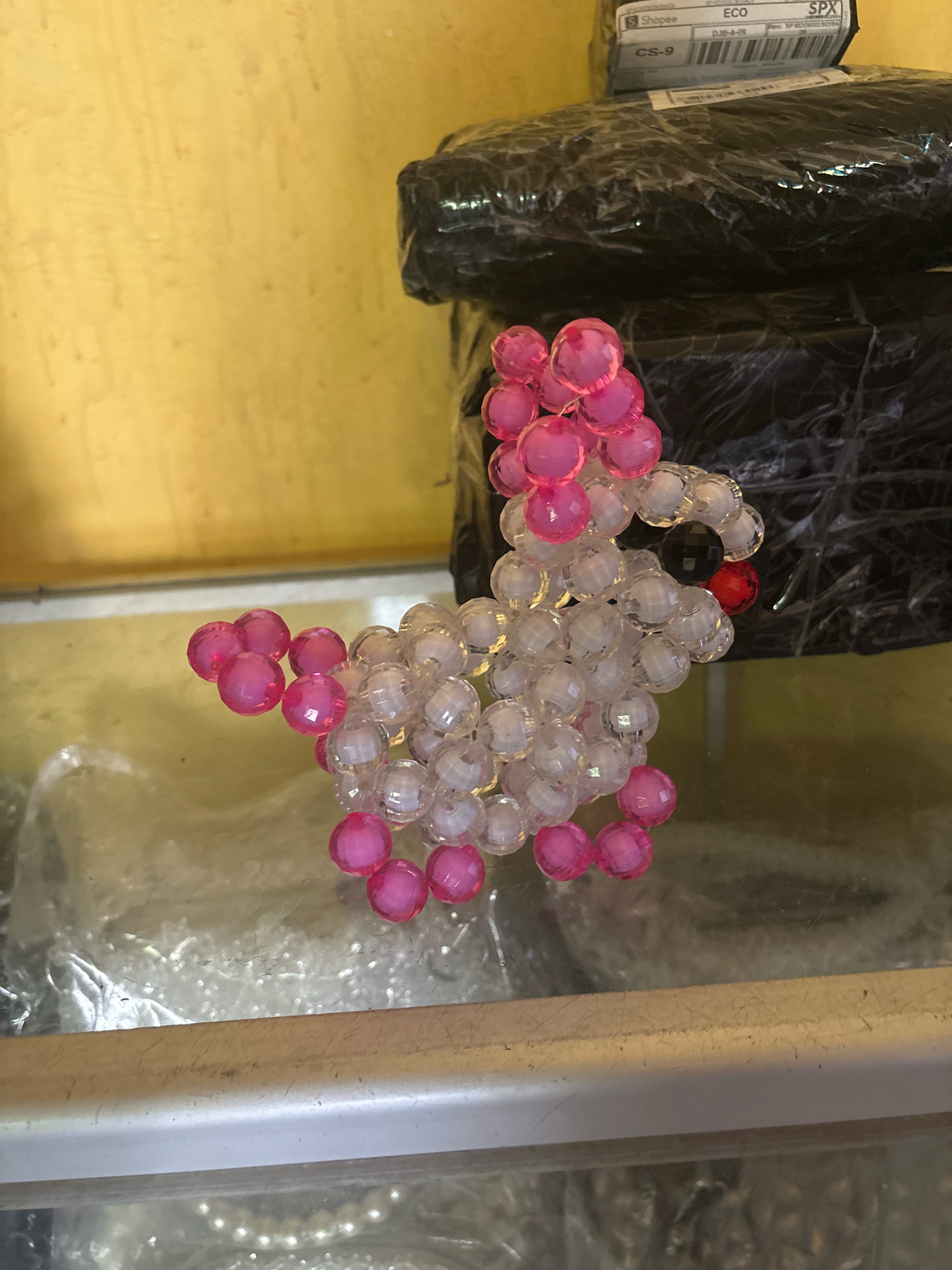 The image shows a close-up of a beaded craft shaped like a chicken, made of pink and clear beads. The chicken is positioned on a glass surface, with a yellow wall in the background. Behind the chicken, there are black plastic-wrapped packages stacked, one of which has a shipping label with "Shopee" visible. This scene is likely from Jambi, Indonesia, as evidenced by the "Shopee" label, a popular e-commerce platform in Southeast Asia, and the overall aesthetic which is consistent with typical retail or storage environments in the region. There are no people or animals present in the image.