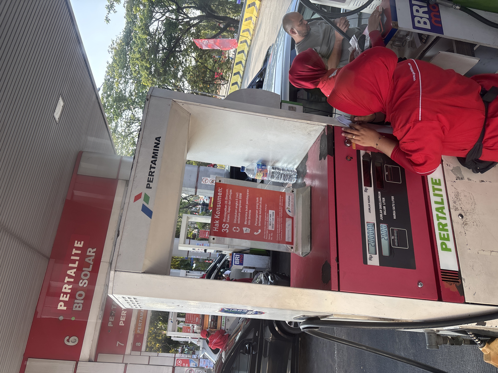 An outdoor shot captures a Pertamina gas station in Karawang, Indonesia. The focus is on a red and white fuel dispenser with digital displays. One display shows "200,000 Rp" for "JUMLAH HARGA" (total price) and "20.00" for "JUMLAH DIKELUARKAN DALAM LITER" (volume dispensed in liters). Below that, "HARGA SATU LITER" (price per liter) is partially obscured, showing "Rp" and "00000". The brand name "PERTALITE" is prominently displayed in green on the lower section of the dispenser.
Two people, likely gas station attendants, are visible. One, wearing a red uniform and headscarf, is attending to a customer in a car, their back is to the camera. Another individual, a male customer, is visible in the car, looking down at papers.
In the background, a white car is being refueled by another attendant in a red uniform. Trees and a blue sky suggest it is daytime. A red banner with a white logo is visible further back, along with other gas pumps and vehicles. The overall scene depicts a typical transaction at an Indonesian fuel station.