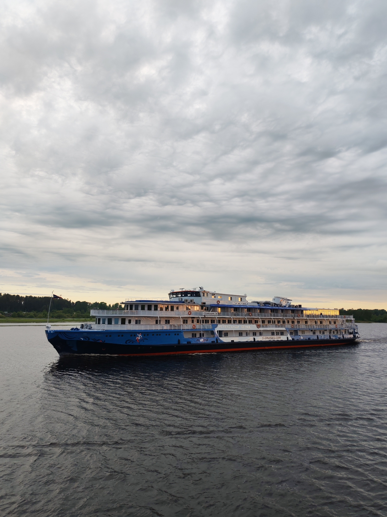 A long, multi-story river cruise ship is moving on a body of water, possibly the Volga River near Myshkin, Russia. The ship has a blue and white hull with a red stripe near the waterline. It is carrying passengers and crew. It appears to be dusk or dawn, as evidenced by the light in the sky and reflections in the water. The sky is mostly overcast. There is land visible in the background. The water is dark and slightly choppy. The ship's name, "Александр Бенуа", and the website "S-CRUISES.COM" are visible on the side.