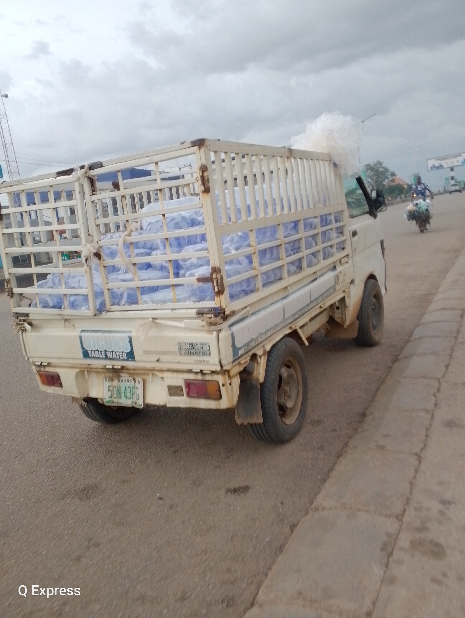 A white truck is transporting a large quantity of bottled water. The truck's bed is enclosed by a white metal cage, and it is filled with rows of blue-bottled water, mostly obscured by plastic wrap. A white material, possibly more plastic wrap or bags, is piled up on the back of the truck, extending above the cage. The truck has a license plate that reads "5ON-43RG." A sticker on the back of the truck says "VIGONS TABLE WATER."
In the background, a person on a motorcycle is seen riding away from the camera, carrying a large load of items. The scene appears to be outdoors, likely on a road, with a paved surface and a concrete curb. The sky is overcast, suggesting a cloudy or possibly rainy day. The lighting is diffused. The overall impression is of a delivery or transportation activity. The visible text confirms the product being transported is "VIGONS TABLE WATER." The image is watermarked with "Q Express" at the bottom left corner.