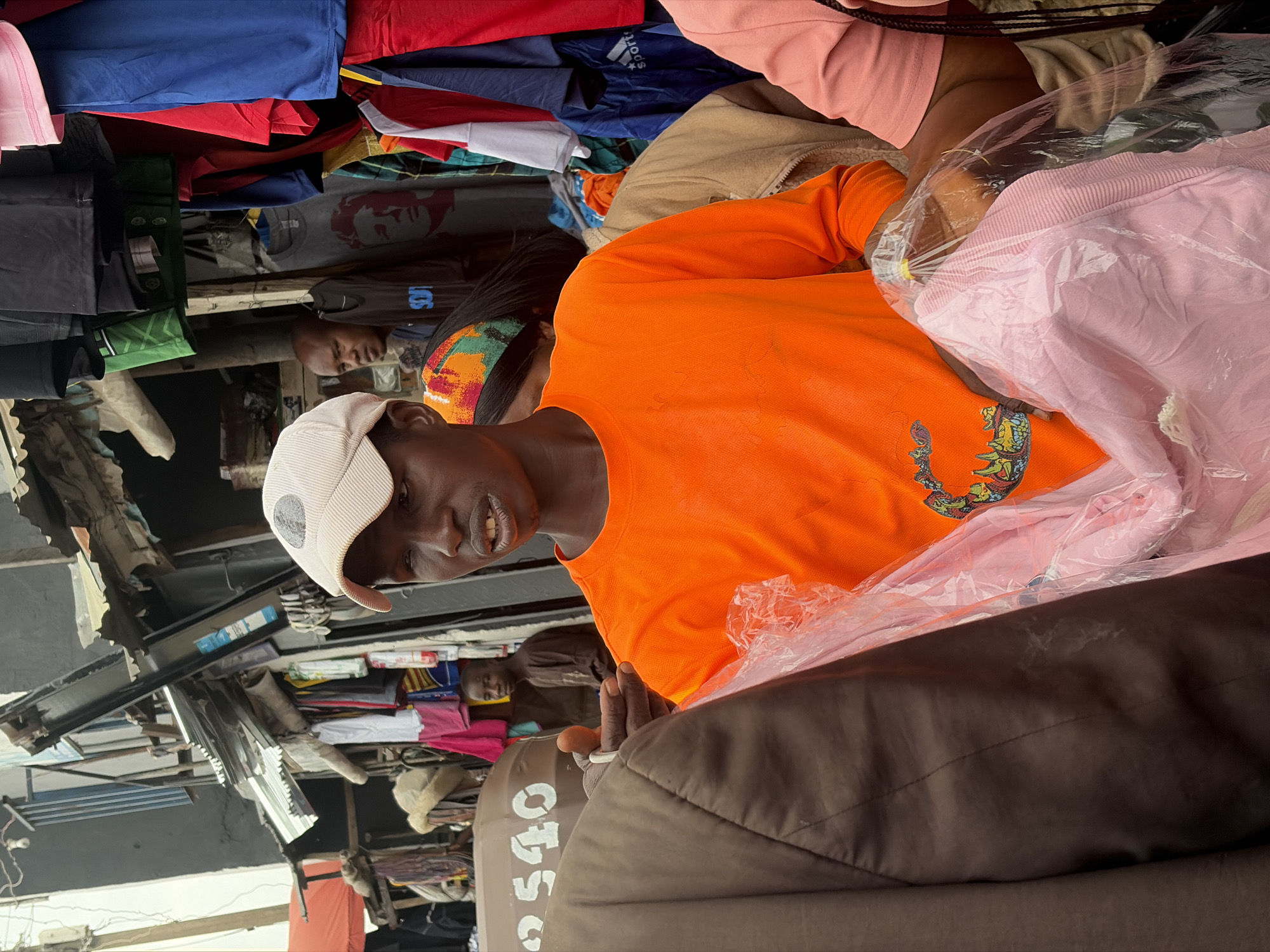 In Bukuru, Nigeria, a male vendor wearing an orange long-sleeved shirt and a beige baseball cap stands in an outdoor market. He is holding a clear plastic bag filled with pink clothing. To his left, another person, partially visible, wears a brown jacket. Behind the vendor, various clothing items hang from stalls, and other people are present in the background. The market appears to be an open-air setting with stalls constructed from corrugated metal and wood.