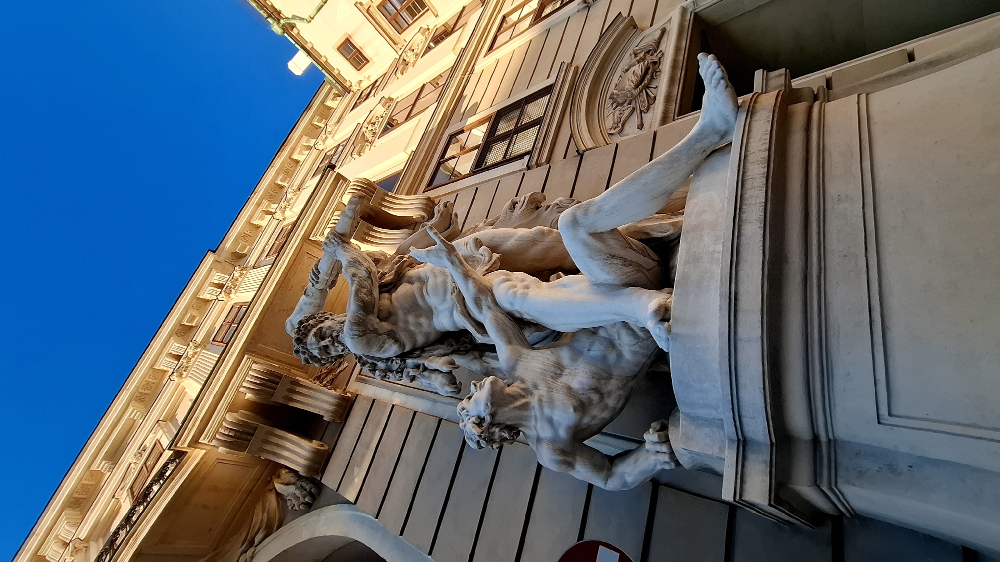 The image shows a low-angle, close-up shot of a white marble sculpture depicting two male figures wrestling. The sculpture is positioned on a pedestal in front of a building. The building appears to be made of light-colored stone and features classical architectural elements such as columns, cornices, and windows with dark frames. The sky above is a clear, bright blue. The sculpture is illuminated by warm, directional light, suggesting it is either sunrise or sunset.
The foreground sculpture features two muscular male figures. One figure is in a kneeling or seated position, looking upwards with an outstretched arm. The other figure is standing and appears to be overpowering the kneeling figure, with one arm raised as if to strike. Both figures are nude and sculpted with detailed anatomy.
The building in the background is multi-story with visible windows. Some of the windows have grates. The architectural style suggests it is a historic building. The overall impression is one of classical grandeur and artistic mastery. This sculpture is likely part of the exterior decoration of a significant building. Based on the architectural style and the quality of the sculpture, it is highly probable that this image was taken in Vienna, Austria, likely at a location like the Hofburg Palace or another imperial building.