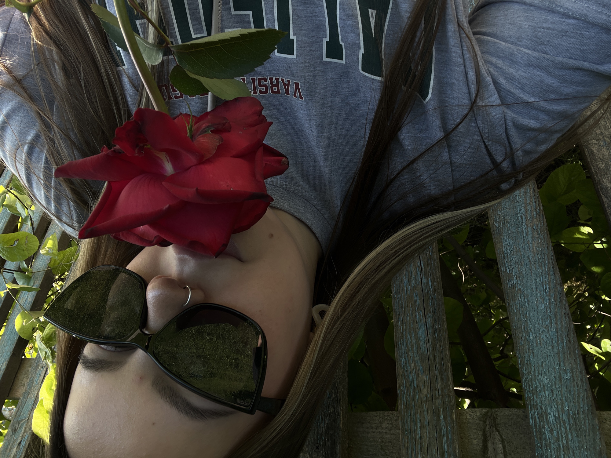 A young woman is depicted outdoors in Novopokrovskaya, Russia, holding a vibrant red rose to her face, positioned as if smelling it. She wears large, black, cat-eye style sunglasses that reflect green foliage and possibly blue sky. A silver hoop nose ring is visible in her right nostril, and a light-colored, irregularly shaped earring is in her left ear. Her long, light brown hair with highlights frames her face and extends over her shoulders. She is dressed in a grey crew-neck t-shirt featuring partially visible maroon text that reads "VARSITY" above larger, dark green letters. The background consists of a weathered wooden fence or trellis with peeling blue-green paint, behind which natural green foliage, trees, and sunlit areas are visible.