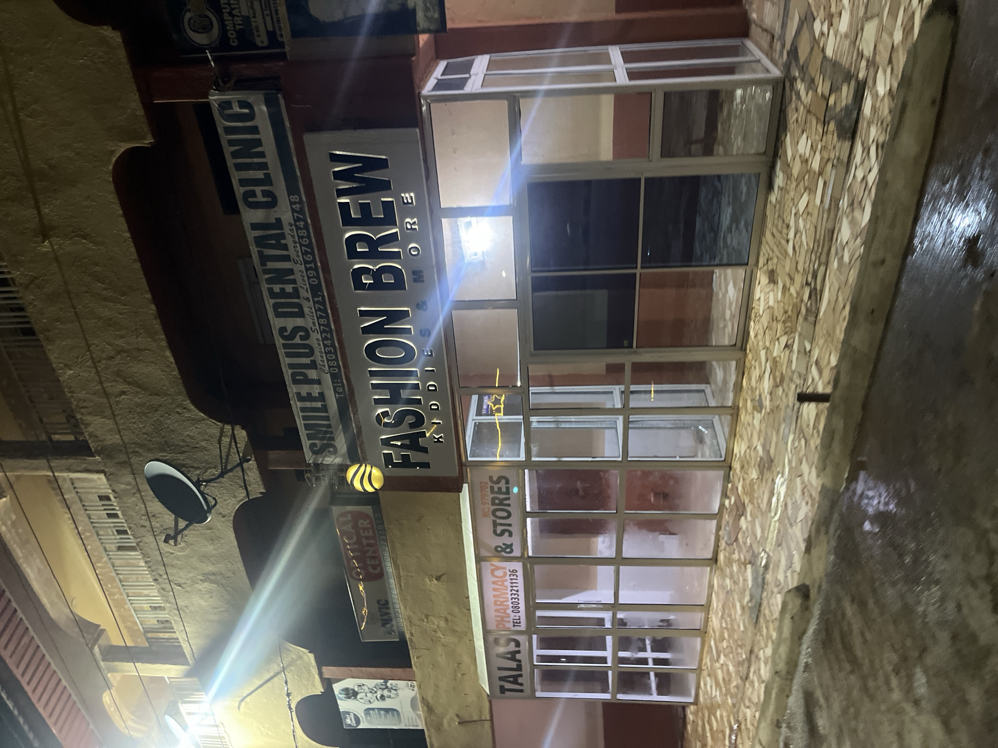 The image captures a storefront in Bukuru, Nigeria, at night. The facade is illuminated by streetlights and internal shop lighting, creating reflections on the wet pavement and the glass storefront. The building appears to be multi-story with balconies and is constructed with stucco and concrete.
Several signs are visible on the building. Above the main storefront, a large sign reads "FASHION BREW KIDDIES & MORE." To the left of this, a sign advertises "OPTICAL CENTER." Further to the left, a sign displays "SOLVIC." Above the "FASHION BREW" sign, another sign reads "SMILE PLUS DENTAL CLINIC" with contact information. To the right of "SMILE PLUS DENTAL CLINIC," a partial sign indicates "COMPUTER TRAIN."
The main storefront is a glass-enclosed space with an aluminum frame. Inside, a sign for "TALAS PHARMACY & STORES" is visible, along with a phone number. The interior of the shop is dimly lit, with some shelving and displays visible. The pavement in front of the store is wet and appears to be paved with irregular stones. A satellite dish is mounted on the exterior wall of the building. The overall scene suggests a commercial area in an urban setting, likely experiencing recent rainfall given the wet surfaces.