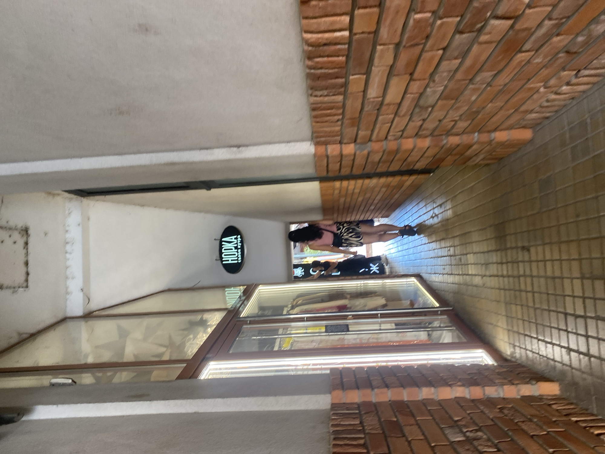 A woman walks down a narrow, covered alleyway. The alley has brick walls on both sides and a tiled floor. On the left side of the alley, there is a storefront with a glass door and windows, displaying clothing on racks. A sign with neon lettering that reads "HOPKA" is visible hanging overhead. The woman is wearing a pink top and a patterned skirt, and she is carrying a bag on her shoulder. She is walking away from the camera and towards the end of the alley, where more shops and possibly an outdoor area are visible. The lighting suggests it is daytime, with sunlight filtering into the alley from openings at either end.