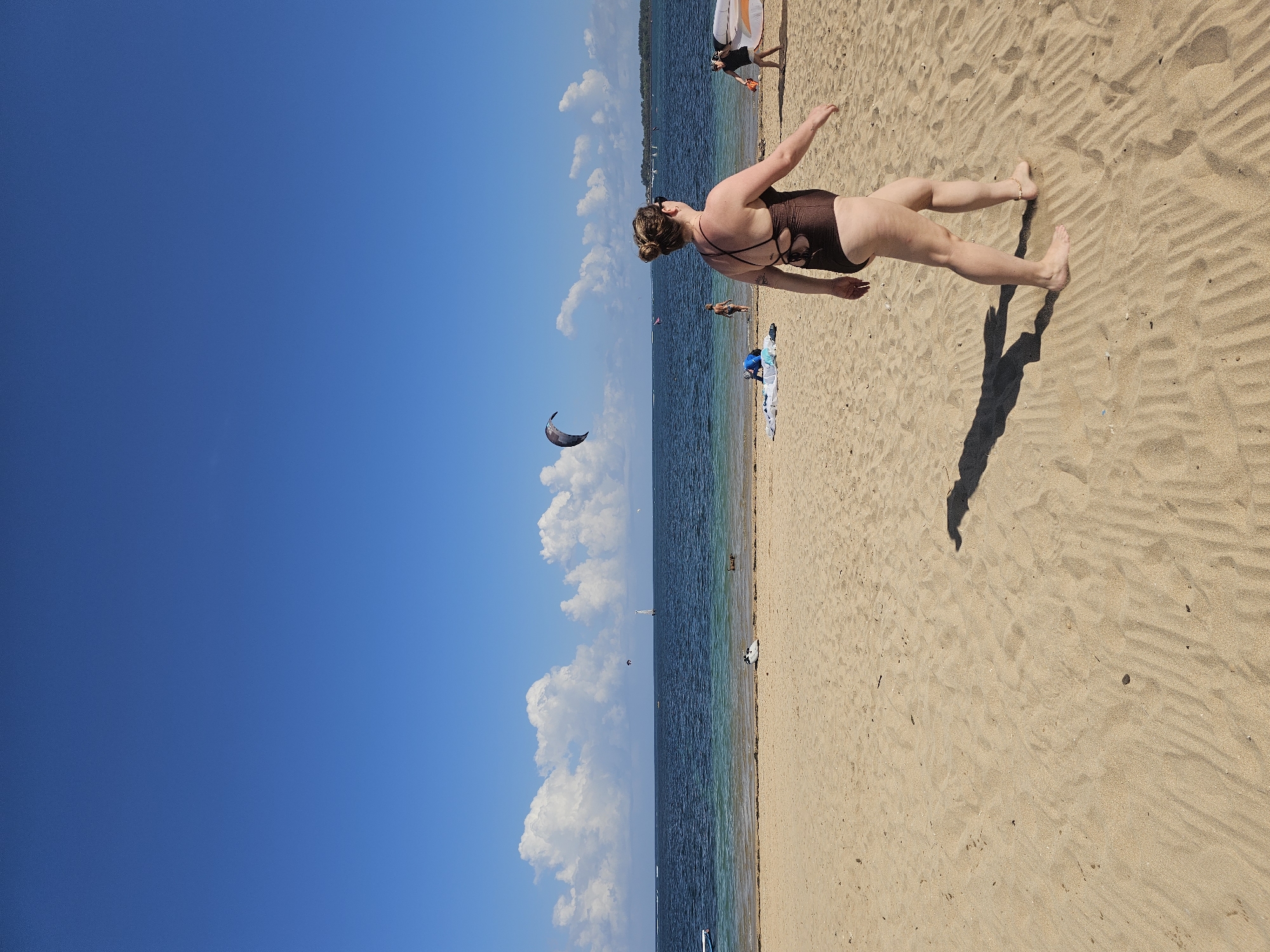 A woman in a brown one-piece swimsuit walks on a sandy beach towards the right of the frame, casting a long shadow to her left. She has her hair pulled up and wears sunglasses. The beach is largely empty, with some scattered beach gear and a few other people in the distance. The ocean is a deep blue with lighter turquoise shallows near the shore. Several kites are visible in the sky above the water, and white clouds dot the bright blue sky. The scene is set on a sunny day at a beach location, possibly in the city of Denpasar, Indonesia, known for its coastal areas and watersports.