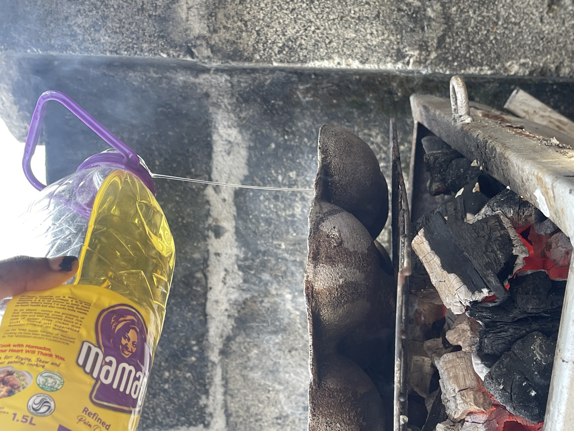 The image shows a cooking setup. A bottle of "mamador" refined palm oil is held in someone's left hand. Next to it is a metal grill with what appears to be grilling stones or rocks, positioned to the left of a charcoal-filled cooking box with glowing embers. The background is a grainy, possibly concrete or stone wall. The environment and the presence of Mamador oil (a cooking oil brand widely available in Nigeria) suggest that this image was taken in Nigeria. Without additional information, pinpointing Ado, Nigeria, as the specific location is not possible.