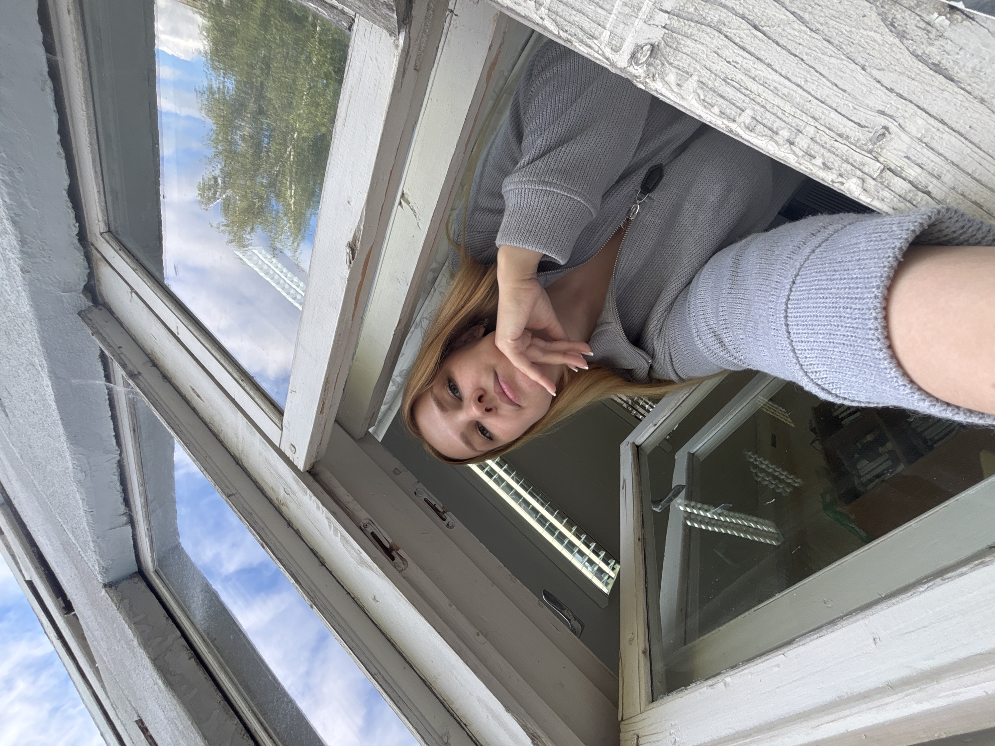 A woman is looking into the camera from a window with an overhead angle. The window frame is painted white and has an aged appearance. The woman has light brown hair and is wearing a gray zippered sweater. Outside the window, there are trees and a cloudy sky. The image was taken in the city of Rusanovo, Russia.