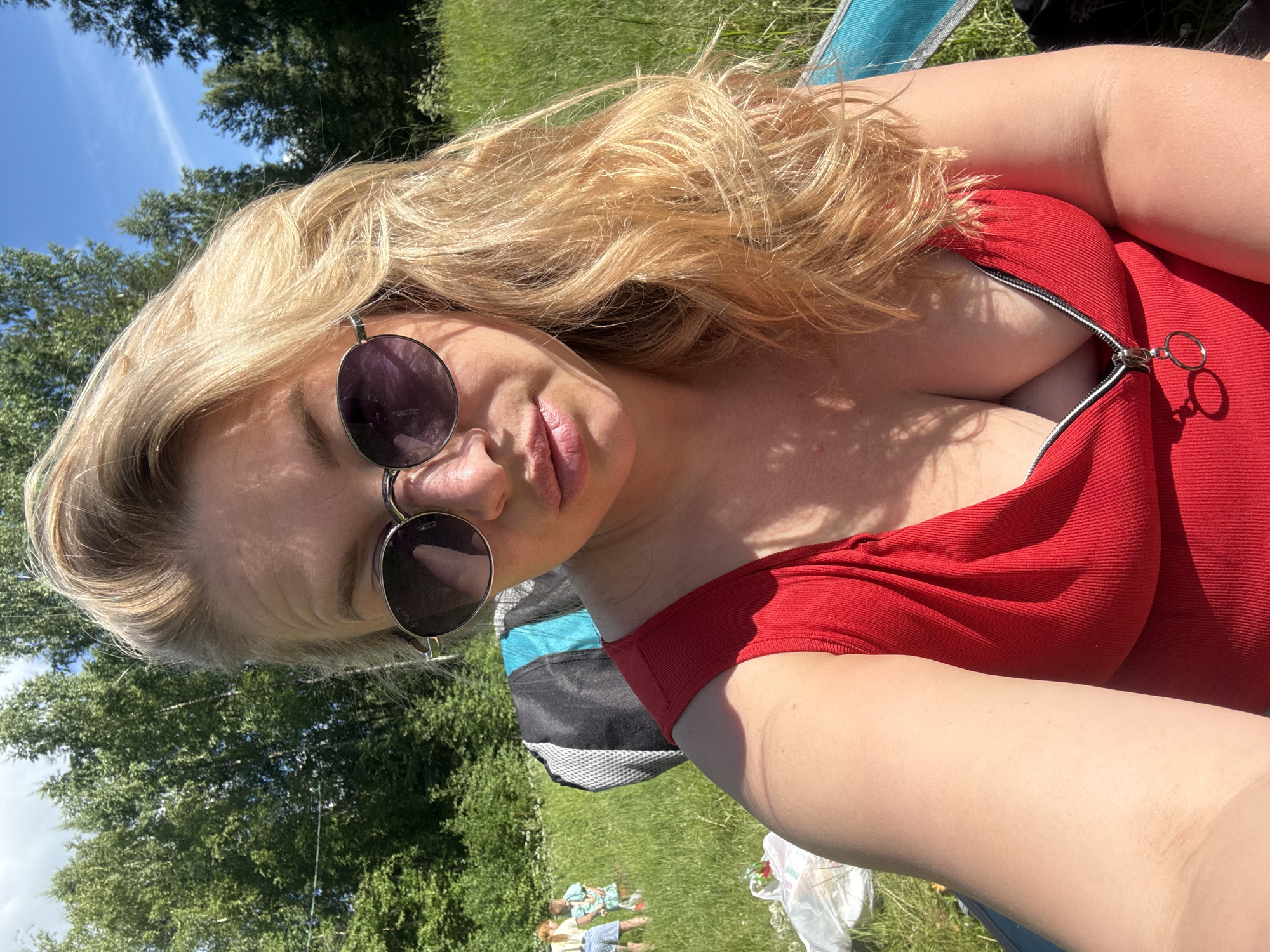 A woman with blonde hair and sunglasses is taking a selfie in a grassy, outdoor setting, likely a park or recreational area, on a sunny day. She is wearing a red top with a zipper. In the background, two young children are visible playing in the grass. The scene appears to be in or near the city of Korotsko, Russia.