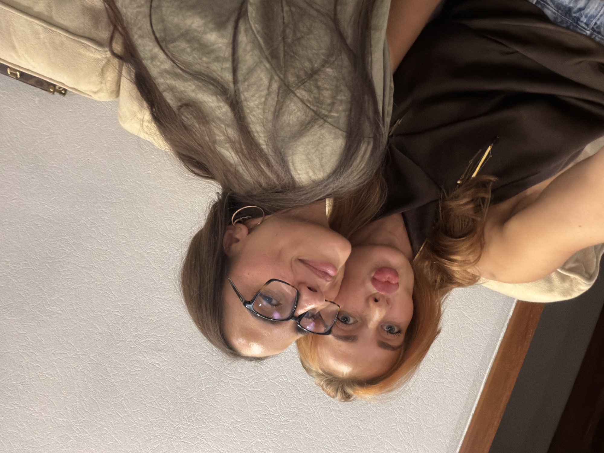 Two women pose closely together in an indoor setting against a white wall. The woman on the left has dark brown hair and wears glasses. The woman on the right has blonde hair and puckers her lips. They are positioned so their faces are near each other. Behind them are portions of a beige couch and clothing in shades of green and brown. The location appears to be in the city of Unknown, Unknown.