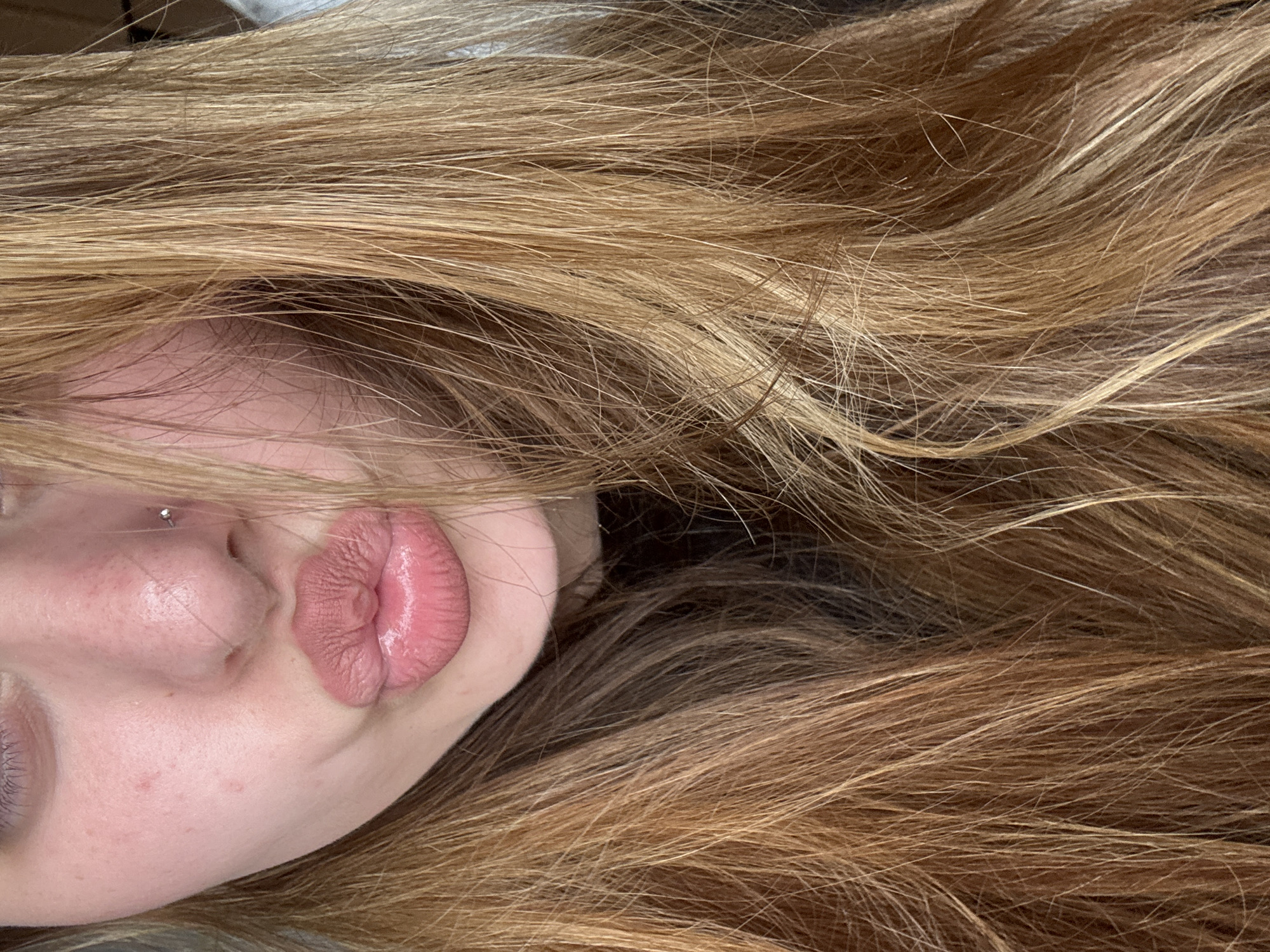 A close-up, tilted portrait features a person with long, wavy, golden blonde and light brown hair that dominates the frame. The person's face is partially visible on the left side, showing their nose, puckered lips, chin, and a sliver of an eye. The lips are coated with a glossy, pinkish-nude lipstick. A small, silver stud nose piercing is visible on the left nostril. There is a hint of light, possibly purple, eye makeup near the visible eye. The skin appears clear with some subtle freckles. The hair cascades across the top, right, and bottom of the image, showing various strands and textures. The overall impression is playful and relaxed, with soft, even lighting suggesting an indoor setting. No text is visible in the image.