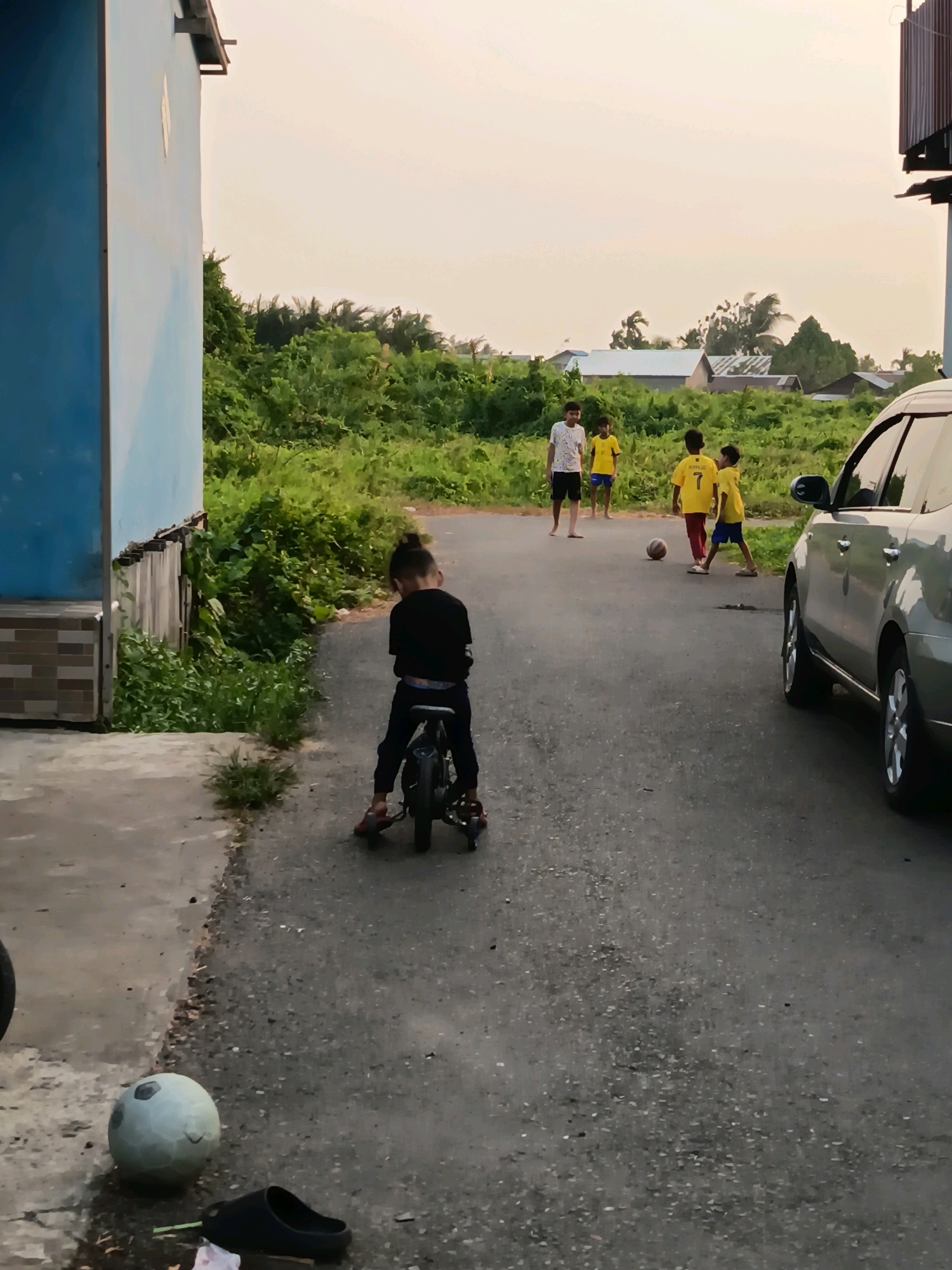 The image depicts a street scene in Banjarmasin, Indonesia, during the late afternoon. In the foreground, a young boy with his hair tied up in a bun is riding a small, dark-colored bicycle with training wheels. He is wearing a black t-shirt and dark pants. The bicycle has visible red accents on the pedals. To the left of the boy, a light blue building with a striped lower facade is visible. The ground is paved with asphalt, with a concrete path on the left. A light-colored ball and a dark sandal are on the ground near the concrete path. Further down the street, four boys are playing with a soccer ball. Two of them are wearing yellow t-shirts, one with the number 7 prominently displayed on the back. The other two boys are wearing a white t-shirt and a yellow t-shirt respectively, along with dark shorts. A silver SUV is parked on the right side of the street, partially blocking the view of the playing children. The background consists of lush green vegetation, several buildings with corrugated metal roofs, and palm trees, characteristic of a semi-rural or suburban area within Banjarmasin. The sky is overcast with a pale yellow hue, suggesting either early morning or late afternoon light.