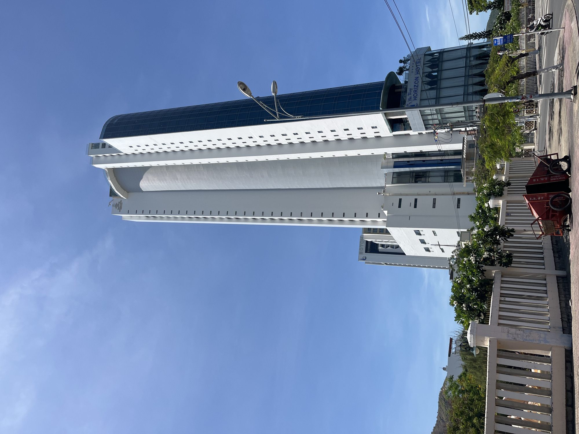 The image depicts the Nha Trang Horizon Hotel in Bac Nha Trang Ward, Nha Trang, Vietnam. The tall, white building has a rounded, glass-enclosed top section. A street runs alongside the building, with a sidewalk, trees, and a white fence. A red utility cart is visible on the sidewalk. The sky is clear and blue.