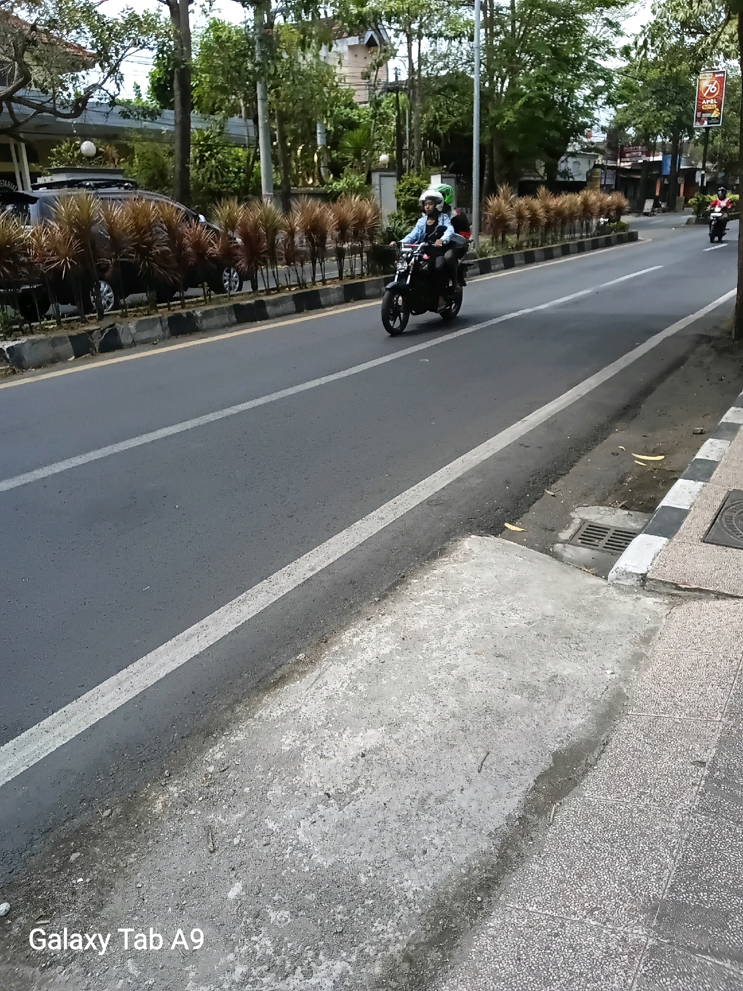 The image depicts a street scene in Kelutan, Indonesia, on a daytime, overcast day. A dark-colored car is parked on the left side of the road, adjacent to a row of decorative plants. The street has two lanes separated by a white line. In the foreground, two individuals are riding a motorcycle. The rider is wearing a light blue shirt and a white helmet, while the passenger has a green helmet. Another motorcycle with a rider wearing a red shirt is visible further down the street, moving in the same direction. Trees line the right side of the street, and a building with a red sign is partially visible in the background. The sidewalk on the right side of the road features a textured surface with a black and white striped curb. A drain grate is present on the sidewalk. The bottom left corner of the image displays a watermark that reads "Galaxy Tab A9".