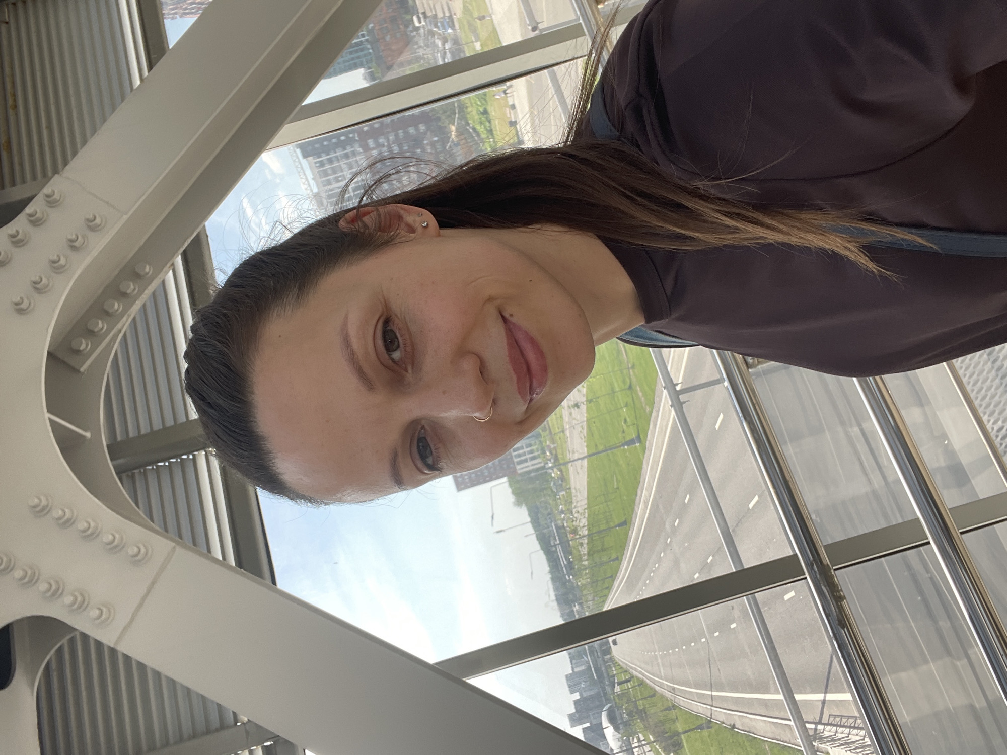 A selfie of a woman with brown hair and brown eyes. She has a nose ring and an earring. The photo appears to be taken indoors, likely on a bridge or elevated walkway, judging from the structure in the foreground and the roadway visible through the windows. The city is partially visible in the background, with buildings and green spaces. The woman is wearing a brown top.