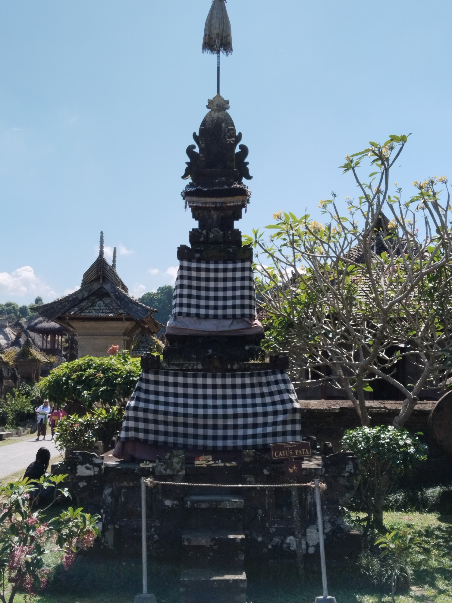 In Bangli, Indonesia, a tall, multi-tiered stone monument, adorned with traditional black and white checkered fabric, stands prominently in an outdoor setting. The monument is capped with an umbrella-like structure and intricate carvings. To the right, a tree with sparse branches and small yellow flowers provides a natural element. In the background, several traditional Balinese buildings with layered roofs are visible, suggesting a temple or cultural site. A paved path runs through the scene, with two individuals in traditional attire walking away from the camera on the left side. A person is crouched near the base of the monument on the left, facing away from the viewer. The environment is sunny with a clear blue sky and scattered white clouds. A small sign with text that appears to read "CATUS PATA" is visible near the base of the monument.