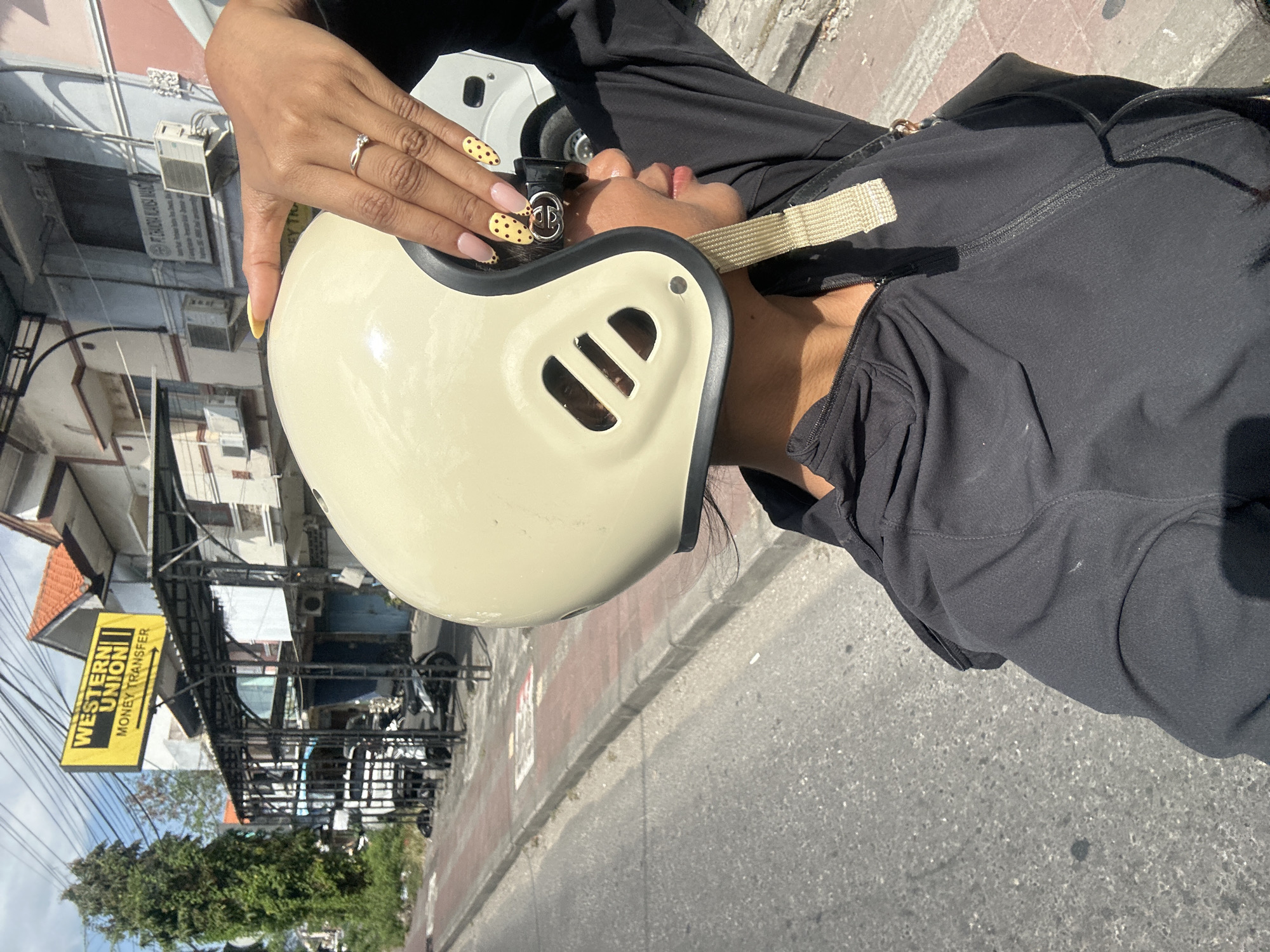 A woman, partially obscured by a cream-colored helmet, is captured in a selfie on a sunny day. She is wearing a black zip-up hoodie and her right hand, adorned with yellow nails decorated with brown polka dots and a diamond ring, rests on top of the helmet. The background reveals a street scene in Denpasar, Indonesia, with a prominent yellow sign for "Western Union Money Transfer" visible above a covered structure. Several motorcycles are parked under the structure, and a white vehicle is partially visible to the right. The street appears to be paved with asphalt, with a red-brick sidewalk lining the road. Buildings with light-colored facades and tiled roofs are visible behind the street. The overall impression is one of an everyday urban environment.