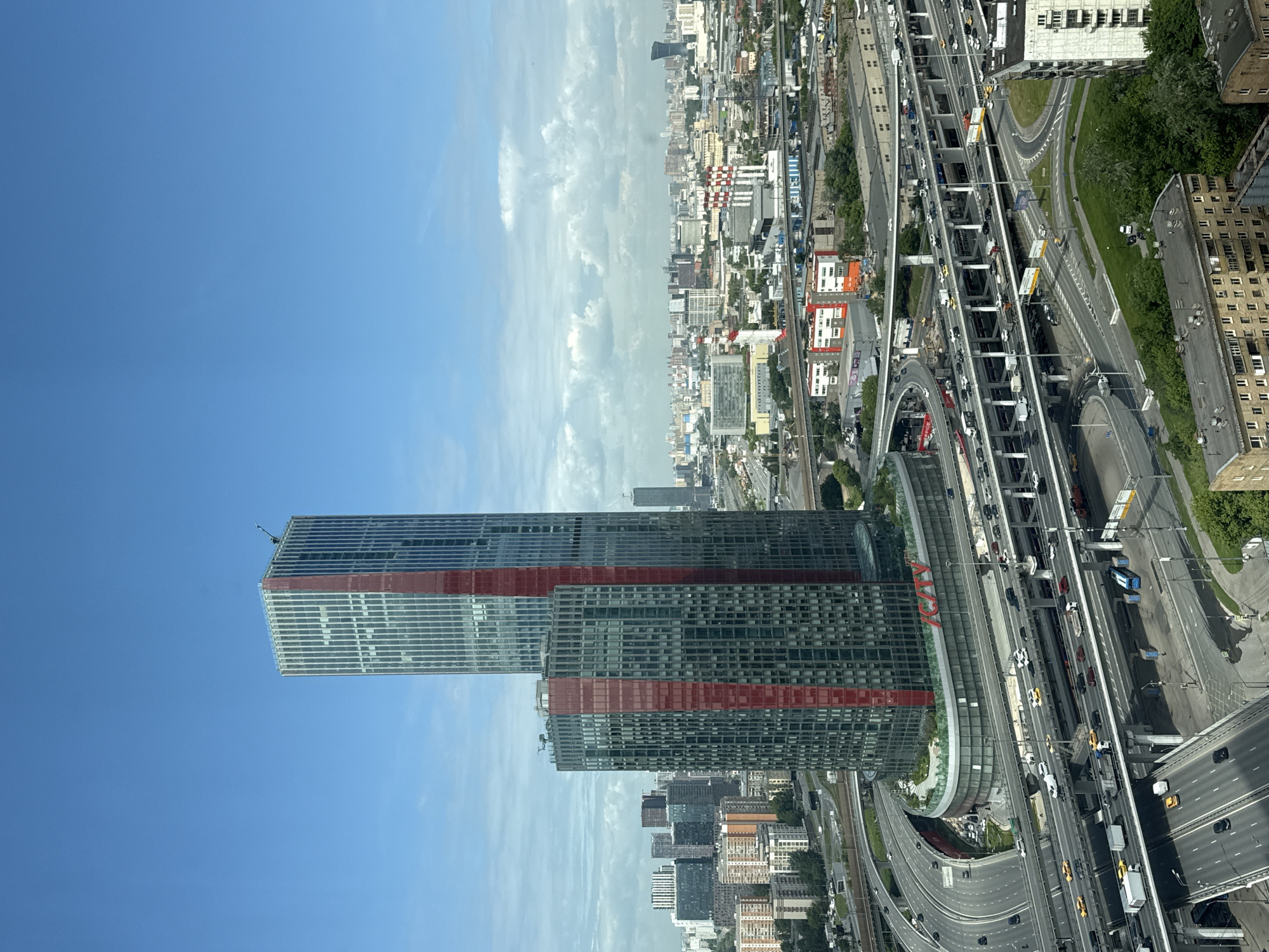 This high-angle, aerial photograph showcases a dynamic urban landscape in Khimki, Russia, under bright daylight.
The central subject is a tall, modern skyscraper with a glass facade, prominently featuring distinctive red horizontal bands. At the base of this building, large red letters spell out "AGAVA". To the right of the skyscraper, a complex system of multi-lane highways and elevated roadways crisscrosses the scene, bustling with active vehicle traffic including numerous cars, trucks, and vans.
Beyond these immediate features, a dense cityscape stretches into the distance, filled with a mix of commercial and residential buildings. The sky above is largely clear blue with some scattered white clouds along the horizon. Patches of green trees and grass are visible alongside the roads and within the urban sprawl. The time of day appears to be midday or early afternoon, characterized by clear weather and good visibility. No individual people are clearly discernible due to the high vantage point.
