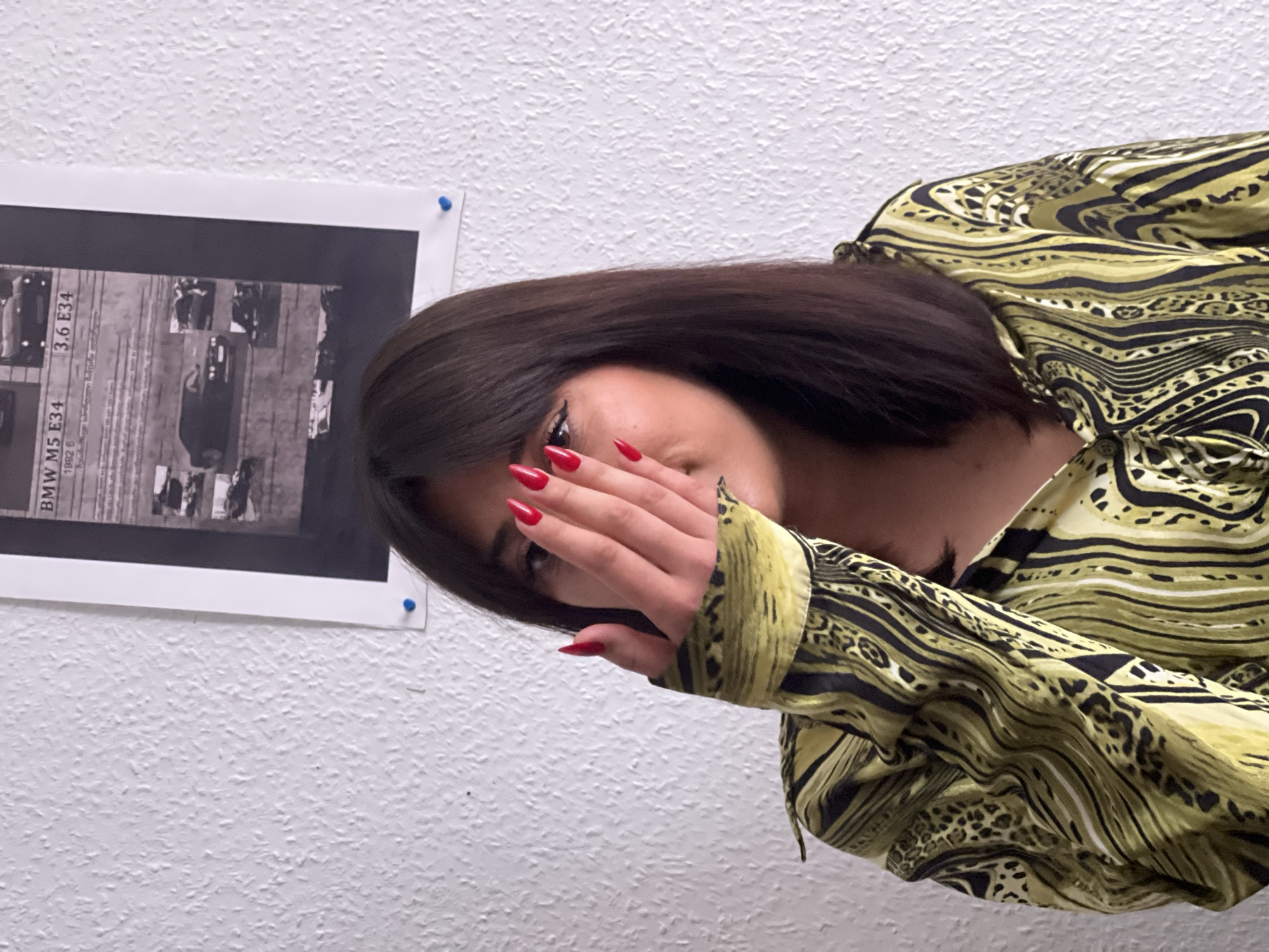 A woman with dark hair and red nails covers part of her face with her hand. She wears a patterned yellow and black shirt. A black and white poster featuring "BMW M5 E34" and "3.6 E34" is affixed to the wall with blue tacks. The background is a textured white wall. This image was not taken in Strasbourg, France.