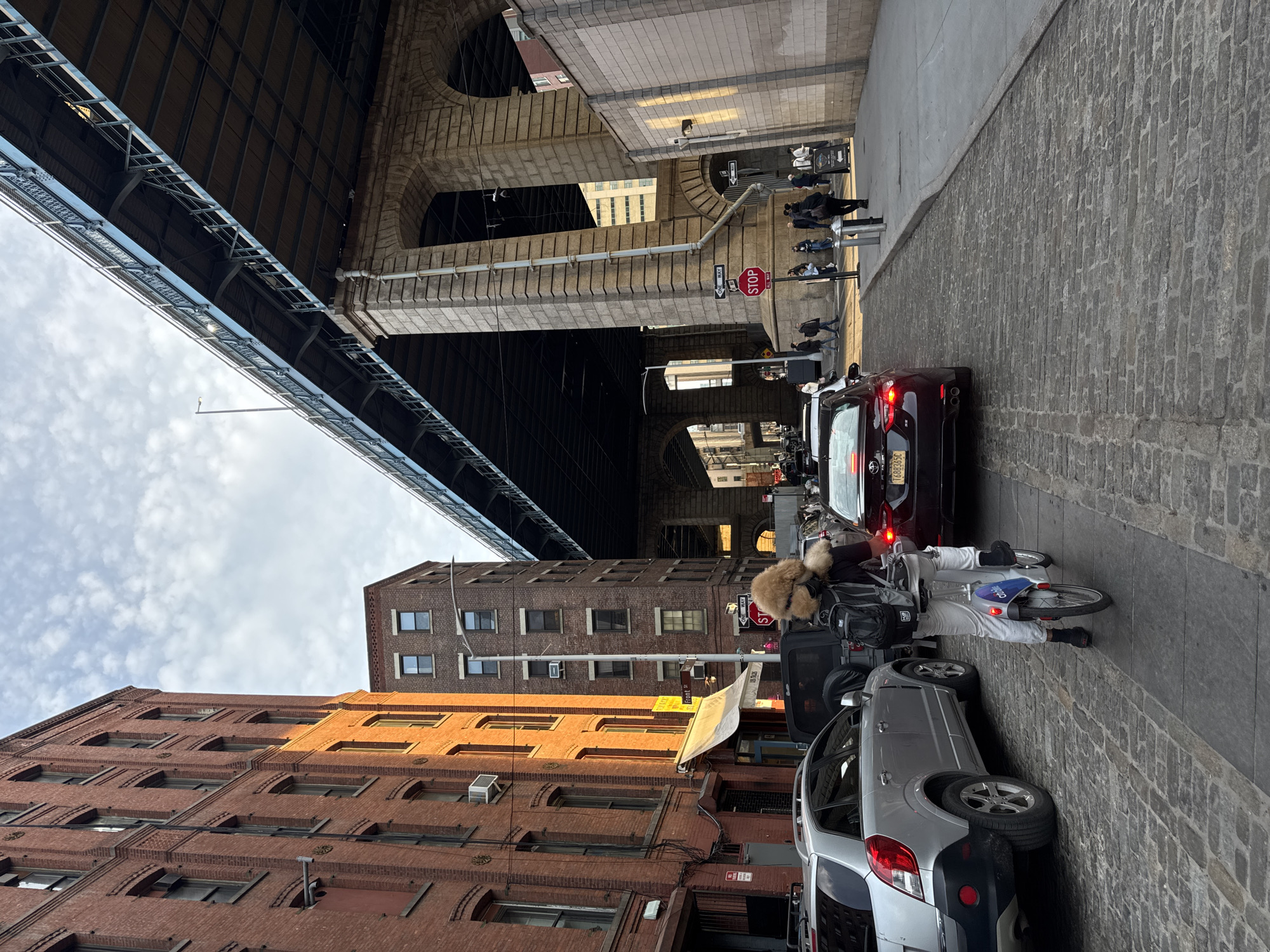 The image shows a street scene in New York City under the Brooklyn Bridge. The view looks up the street with red brick buildings on the left. Cars are parked on the right side of the street, including a silver SUV and a dark-colored sedan. A person wearing white pants, a black backpack, and a furry hat is riding a Citi Bike down the street. The Brooklyn Bridge structure is visible above the buildings and the street. Several pedestrians are visible in the distance, under the bridge. A stop sign is near a brick pillar. The street is paved with cobblestones. The sky is cloudy.