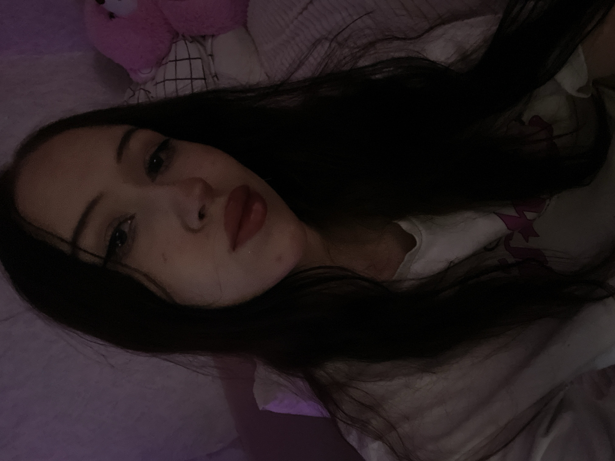 The image is a close-up, dimly lit selfie of a young woman with long, dark hair. She is wearing a white t-shirt. Her face is the primary focus, with her lips slightly parted and her eyes looking towards the camera. A pink stuffed animal and a bedspread are visible in the background, along with a purple wall. The image provides no information about the city of Votkinsk, Russia, or any specific location within it.