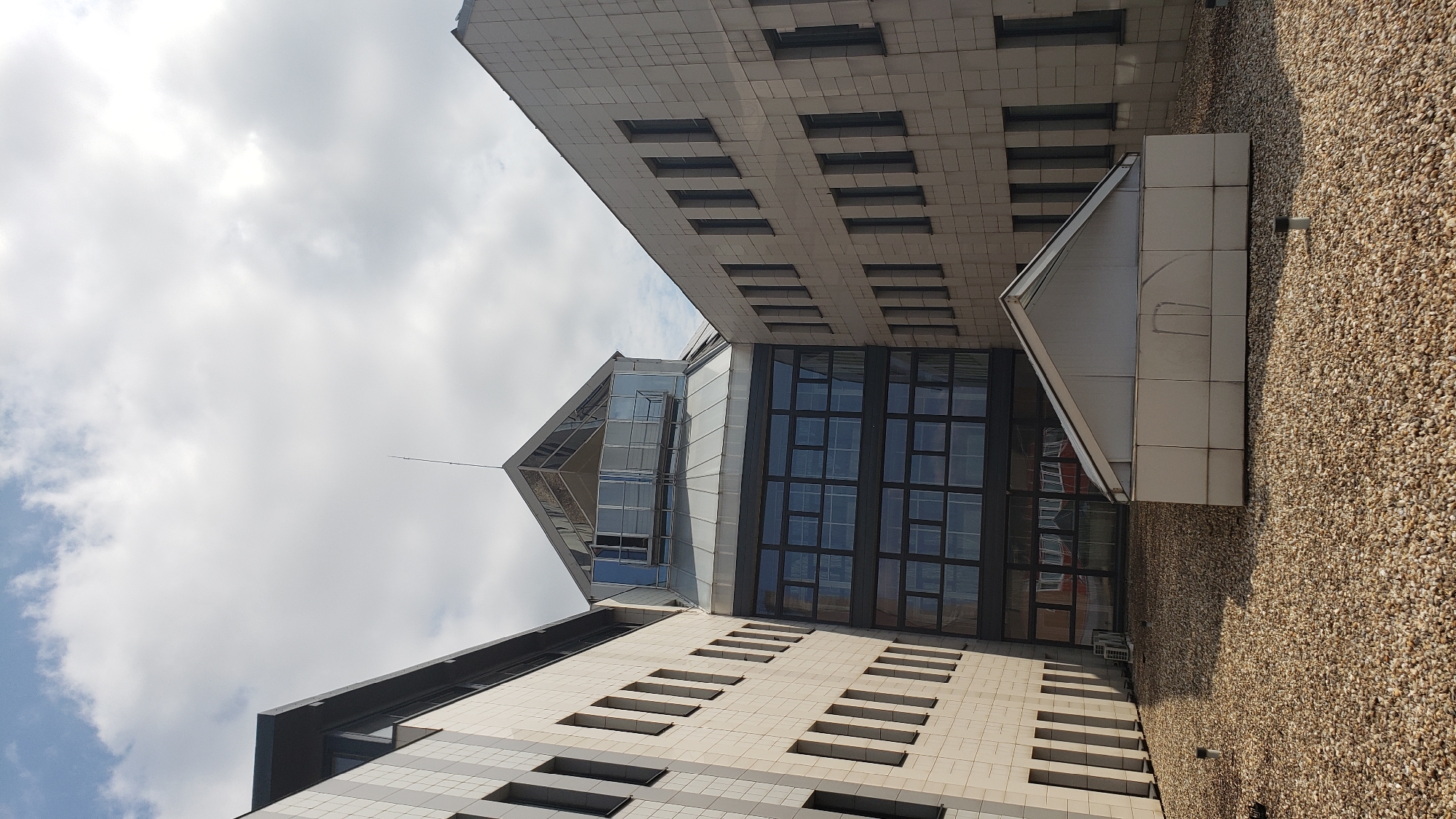 This image depicts a view from a rooftop in the city of Prešov, Slovakia. The rooftop is covered in gravel and features a small, white, gabled structure with a translucent roof. In the background, a modern building with beige tiled facade and numerous windows is visible. A section of the building has a glass-covered atrium with a distinctive angular roof structure. The sky above is partly cloudy, with bright white clouds against a pale blue background. There are no people or discernible actions taking place in the image.