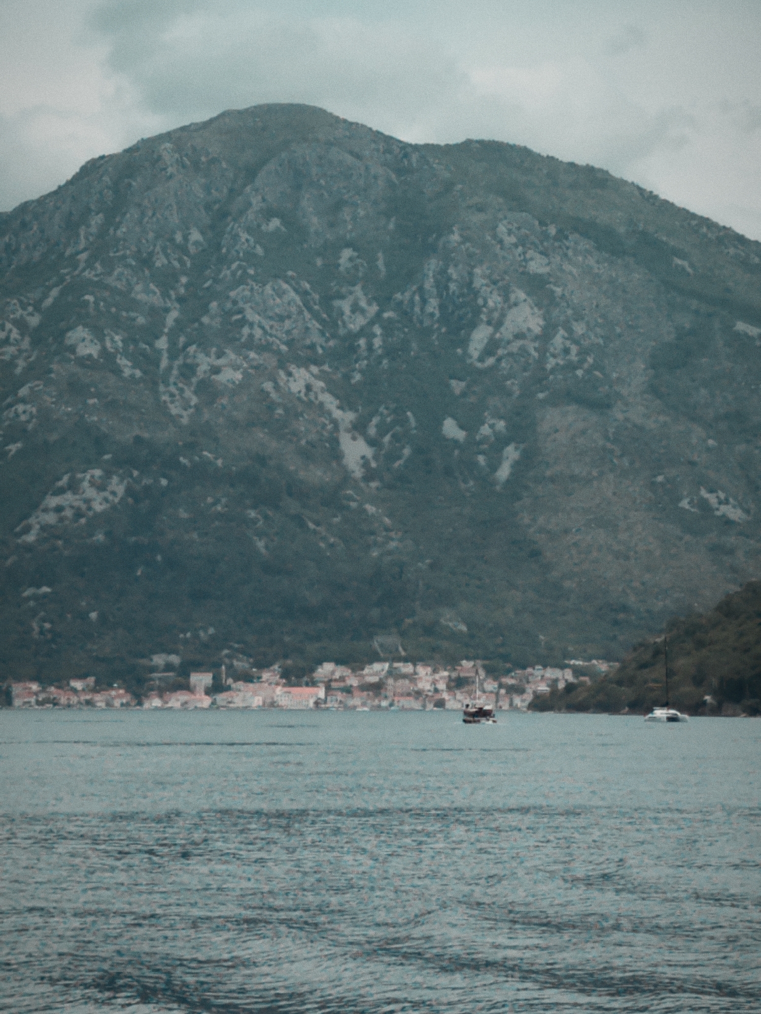 The image depicts a scene in Đurići, Montenegro. A large mountain occupies the upper portion of the frame, exhibiting rocky terrain and sparse vegetation. Below the mountain, a line of buildings representing the settlement of Đurići stretches along the coastline. In the foreground, the water surface displays subtle ripples. Two boats are visible on the water; one is a smaller vessel located closer to the town, and the other a larger, white catamaran positioned farther out. The sky overhead is overcast, suggesting diffused light conditions.