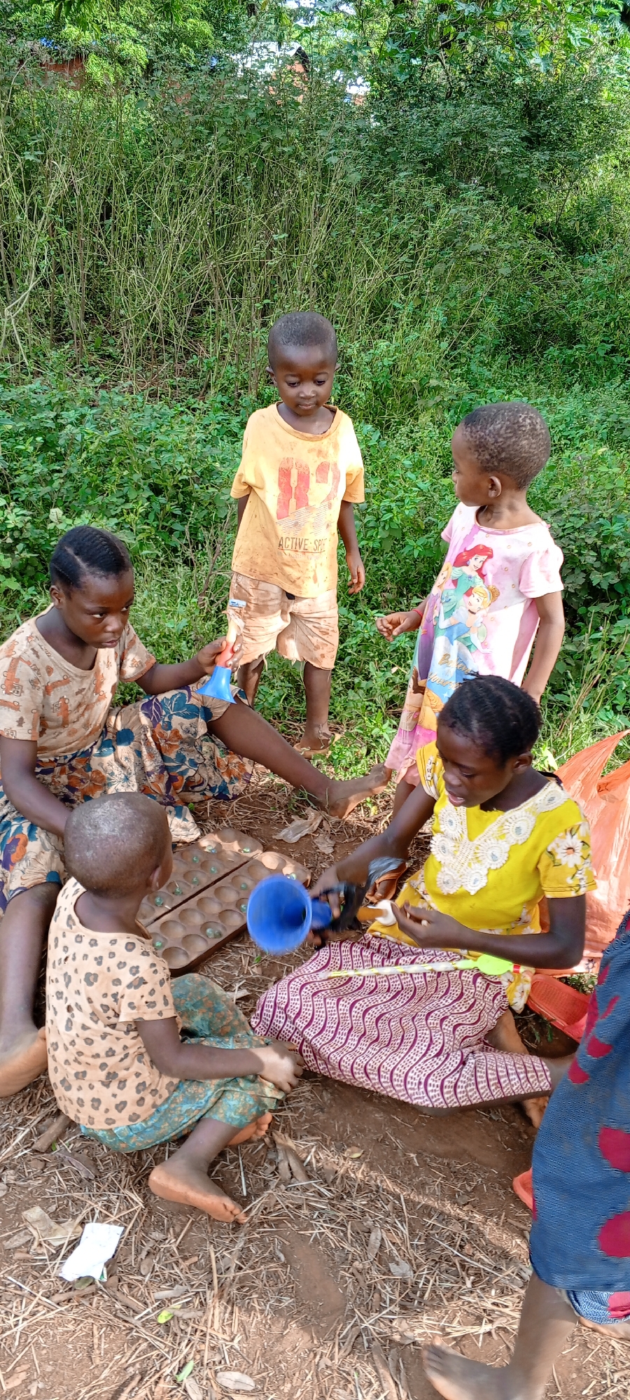 This image captures a group of six children playing outdoors in Ngalula, Democratic Republic of the Congo, during the daytime under sunny conditions. The scene is set on dry, earthy ground mixed with sparse dry grass and leaves, against a dense background of lush green bushes and foliage, suggesting a rural or natural environment.
Four children are seated on the ground, engaged in various forms of play. To the left, a girl in a patterned skirt and top holds a blue plastic item. A wooden mancala board, containing small, light-colored game pieces, rests on the ground between the children. Another child, wearing a spotted top and green patterned pants, is observing the board. To the right, a girl in a bright yellow top with white embroidery and a patterned skirt is actively handling a blue funnel-like object and a yellow plastic stick. Part of a sixth child, wearing a blue and red patterned dress, is visible on the far right.
Standing behind the seated group are two younger children. A boy in a dirty yellow t-shirt with the text "ACTIVE SPE" visible on it and ripped shorts looks downwards, seemingly at the play. Beside him, a girl in a pink dress adorned with Disney princesses looks towards the other children. The children are barefoot and appear focused and calm in their activities. Several orange plastic bags are also visible near the seated children.
