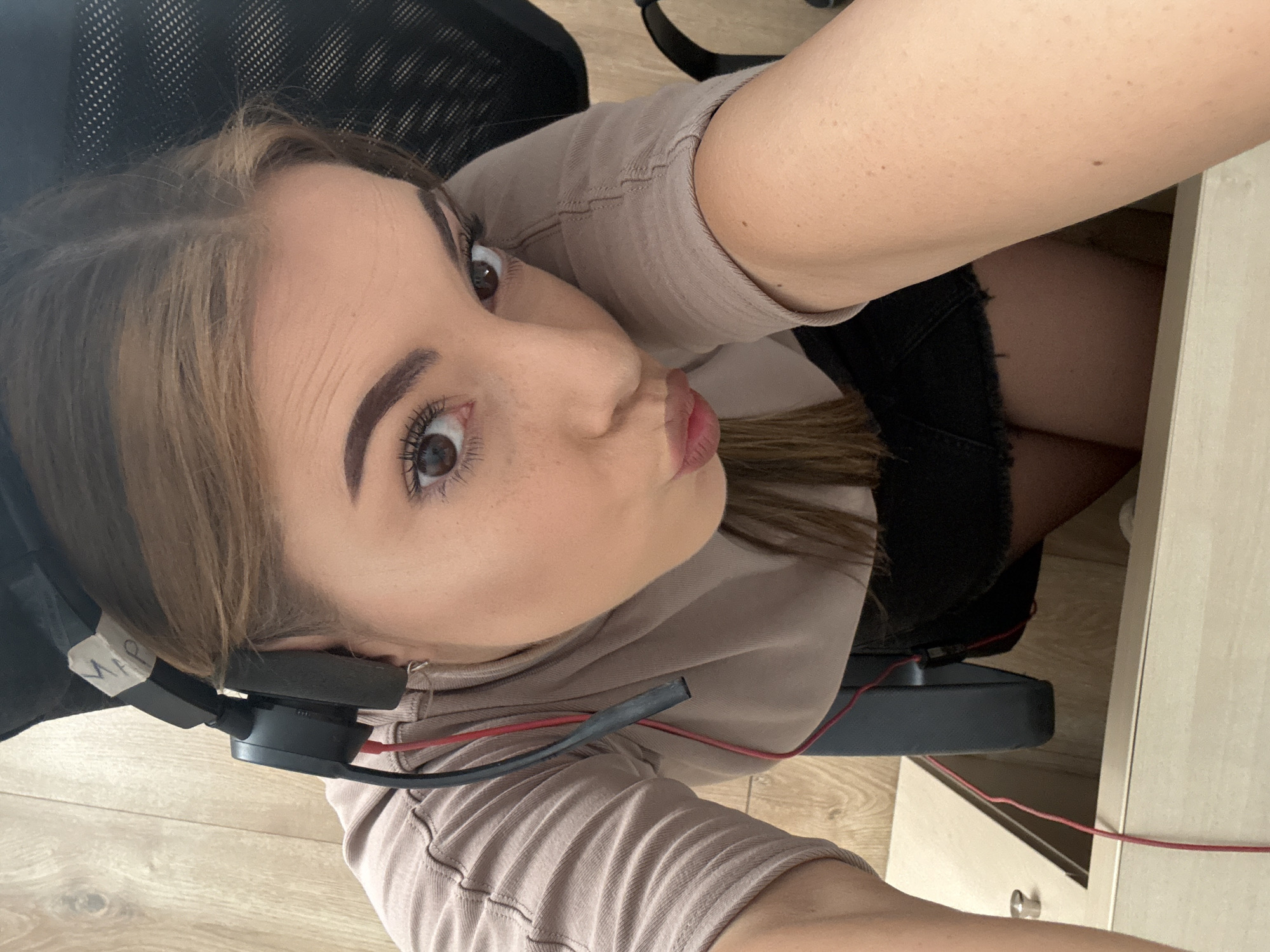 A young female subject is depicted indoors, capturing a high-angle selfie. She has brown hair parted on the side, brown eyes with visible mascara, defined eyebrows, and fair skin with scattered freckles visible on her nose and cheeks. Her lips are pursed in a playful expression. She is wearing a light brown or beige long-sleeved shirt with visible seam details and dark, likely black, frayed-hem shorts. A black over-ear headset with a red audio cable and a black microphone boom is worn on her head. A piece of white tape with the handwritten inscription "MAP" is affixed to the top band of the headset. She is seated, with parts of a black office chair, featuring a mesh back, visible behind her. In the immediate foreground, a light-colored wooden or laminate desk surface is present, with a red cable from the headset running across it and descending into an opening. A metallic fixture or knob is also on the desk surface. The flooring beneath and to the left of the subject is light-toned wooden plank laminate. The scene is located in Sofia, Bulgaria.