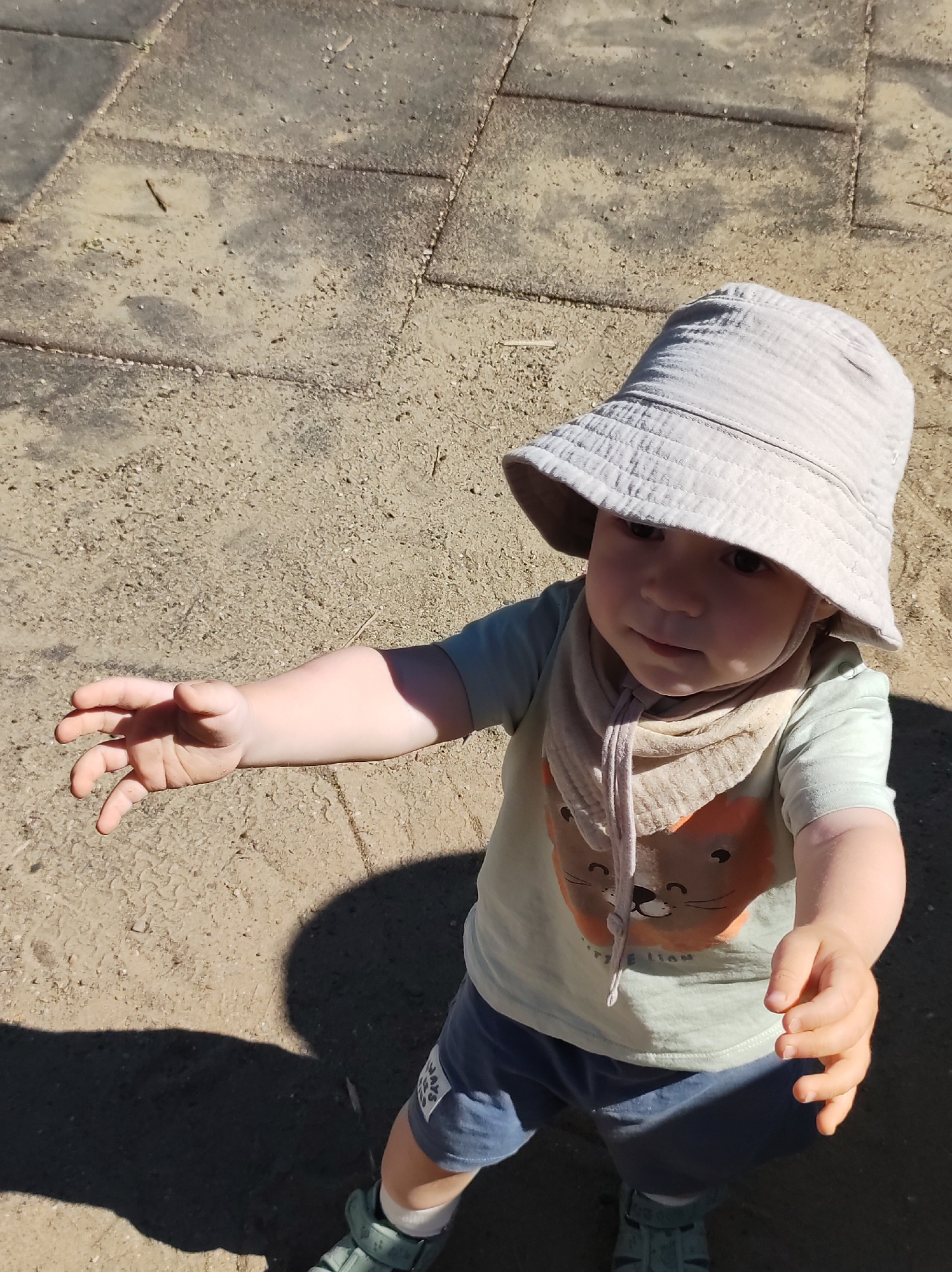 A toddler stands outdoors on a tiled surface, likely in Chaikovsky, Russia, though this is not visually confirmed. The child wears a light-colored bucket hat, a pale green t-shirt with a lion graphic, blue shorts, and green sandals with white socks. They have a beige-colored bandana tied around their neck. The toddler is reaching out with both hands. The ground consists of square grey tiles interspersed with sand. Strong sunlight is casting a shadow of the toddler and their outstretched arms onto the ground.