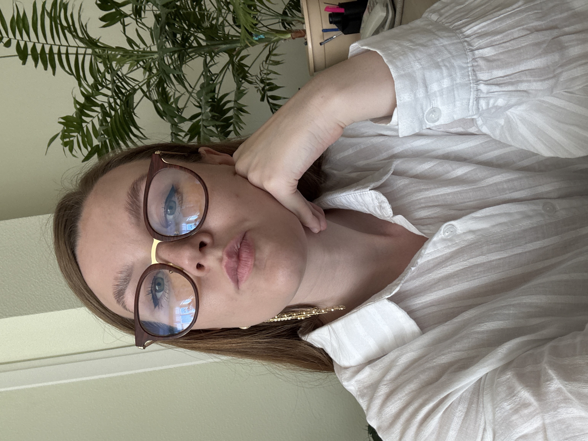 A medium shot, taken from a slightly low angle, captures a young Caucasian woman from the chest up, indoors, with her right hand supporting her chin. She has long, brown hair and is wearing large, brown-rimmed eyeglasses. Her eyes are light blue and she is looking directly at the camera. She has full lips and is wearing a white, collared shirt with vertical pleats. A long, gold earring dangles from her left ear. Behind her to the right is a large potted plant with green, leafy fronds. To the left of the woman is a pale green wall, and to the right, on a desk, are some office supplies, including pens and a phone. The overall lighting suggests an indoor environment during the daytime, likely an office or home office. There is no indication of the city of Izhevsk, Russia, or any other specific location.