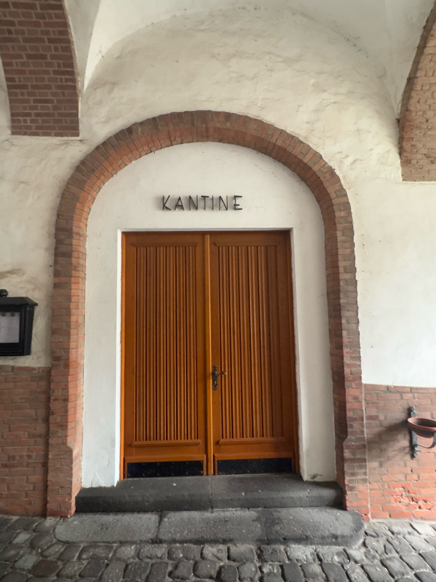 The image shows a brick archway over a white wall, with the word "KANTINE" in black lettering above a set of double wooden doors. The doors are made of vertical planks and have a single black handle. To the left of the doors is a black rectangular object mounted on the wall. To the right, a portion of a brick wall is visible, with a dark object attached to it. The ground in front of the doors is paved with cobblestones and has a stone step. The scene appears to be in the city of Cleves, Germany, specifically in front of a building that houses a canteen.