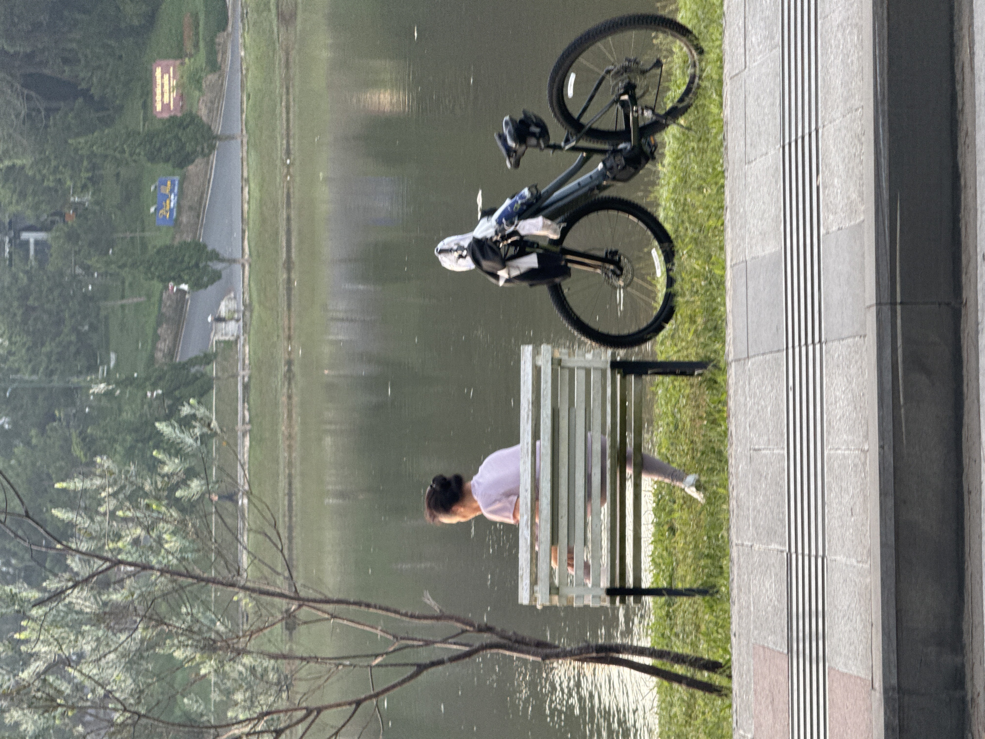 The image depicts Xuan Huong Lake in Da Lat, Vietnam. A person, identified as an adult female by her hairstyle and attire, is seated on a white park bench. She is wearing a pink t-shirt and light-colored pants, and her hair is pulled back in a bun. Her legs are extended forward, and her feet are on the ground. To the right of the bench, a dark-colored bicycle is parked upright, with a white cloth and a water bottle attached to the handlebars. The bicycle's wheels are resting on the grass. In the background, the calm surface of Xuan Huong Lake reflects the surrounding trees and overcast sky. A tree with bare branches stands to the left of the bench. Beyond the lake, a gentle slope is covered with green grass and trees, with a road and a few signs visible on the upper portion of the hill. The foreground shows a paved walkway with tactile paving.