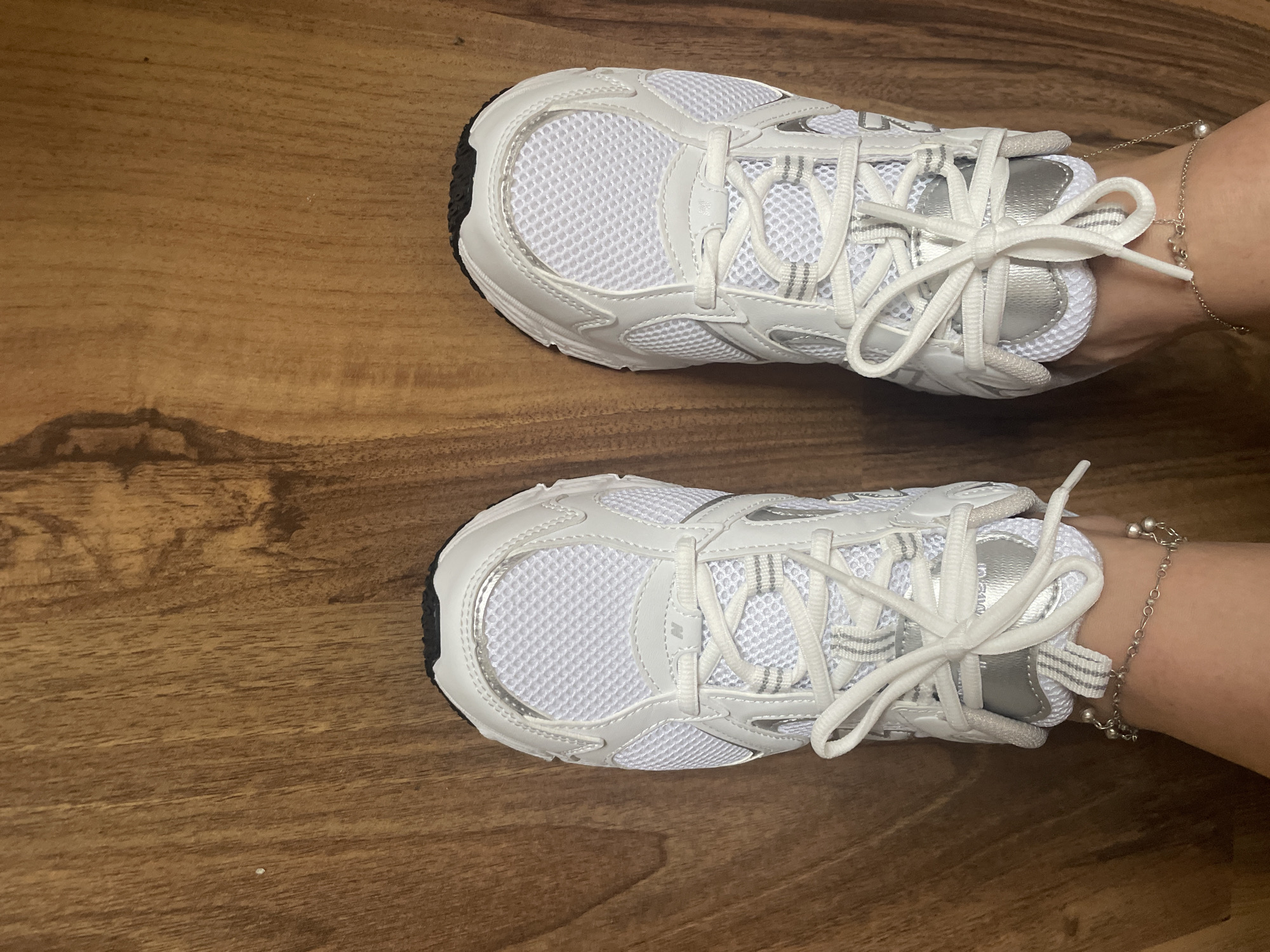 A pair of white athletic shoes with silver accents and white laces are positioned on a wooden floor. Each shoe has a thin silver chain with small charms worn around the ankle. The image is a close-up, top-down view of the footwear. The location is not discernible from the image.