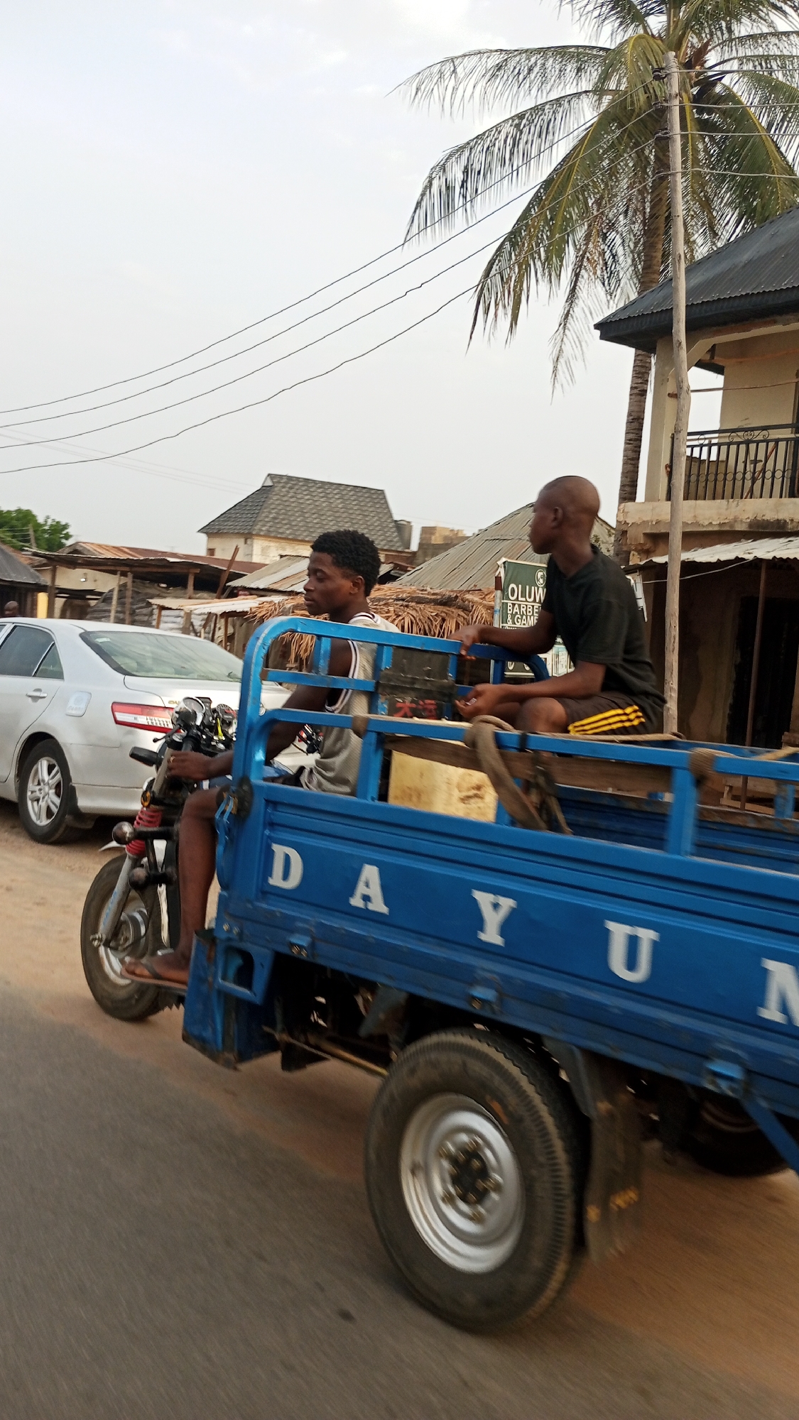 The image shows a blue three-wheeled vehicle, possibly a "Keke Napep" or "tuk-tuk," in Zango-ndaji, Nigeria. Two young men are on the vehicle. One man drives the vehicle and the other sits in the back of the vehicle. The vehicle has the name "DAYUM" on the side. A silver car is parked on the side of the road, and there are buildings in the background with various roofing materials. A palm tree is also visible in the scene. Electrical wires run overhead.