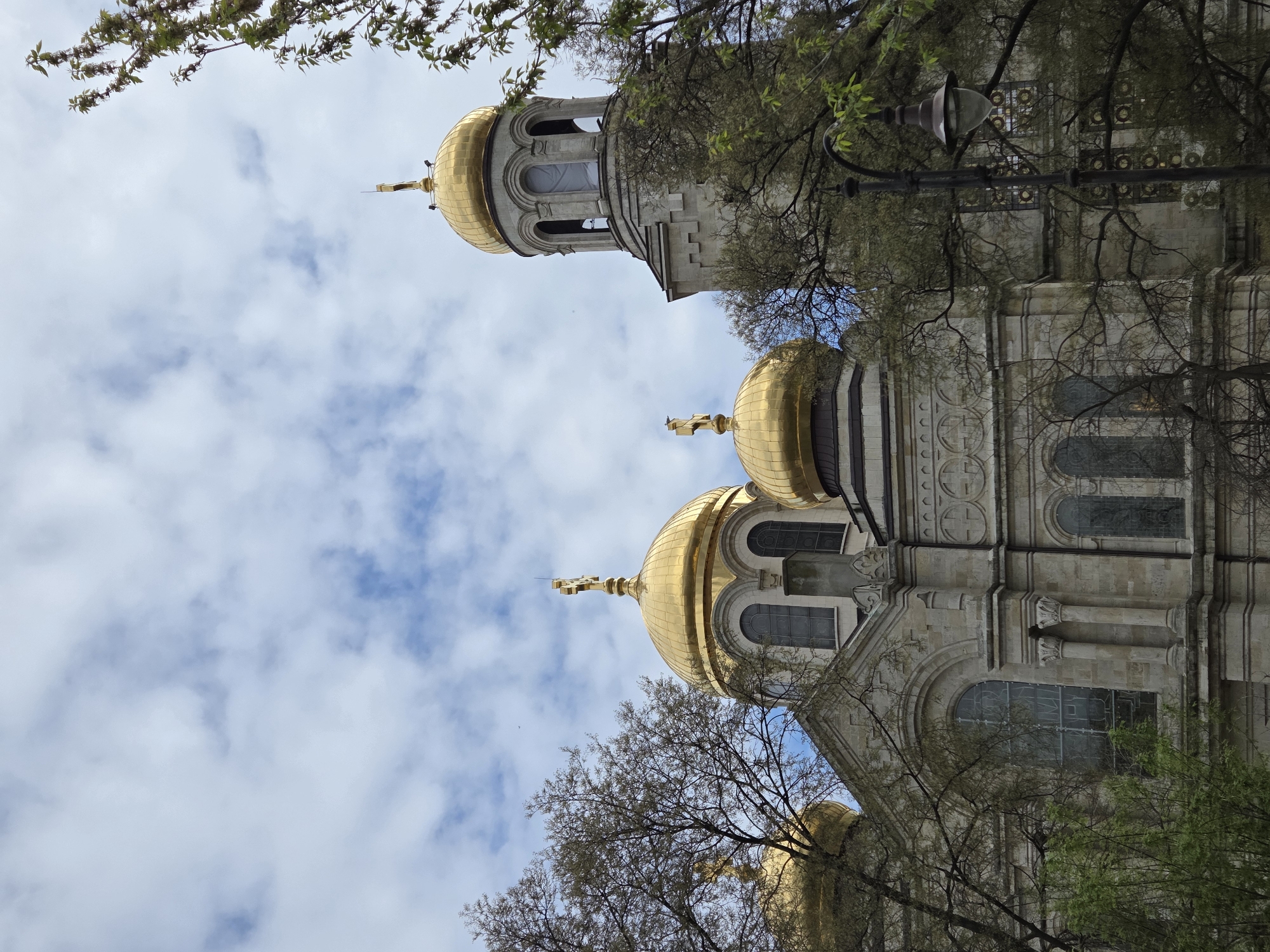 A large, ornate Eastern Orthodox cathedral with prominent golden domes and bell towers dominates the scene, viewed from a low angle looking upwards. The main building features light-colored stone construction with intricate architectural details, including decorative carvings and patterns on its facade, and multiple arched windows.
Several golden domes, each topped with a cross, are visible across the structure. A tall bell tower with arched openings is visible on the right side of the main building. The cathedral is partially framed and obscured by the branches and green leaves of trees, particularly in the foreground and along the upper and side edges of the image.
The sky above is partly cloudy, displaying a mix of blue patches and white clouds, indicating daytime. The natural lighting suggests either morning or afternoon. No specific activities or people are discernible. A street lamp fixture can be observed nestled among the tree branches in one of the cropped views, further indicating an urban or park-like setting around the cathedral. There is no visible text. Given the location context, the building is the Dormition of the Theotokos Cathedral in Varna, Bulgaria.