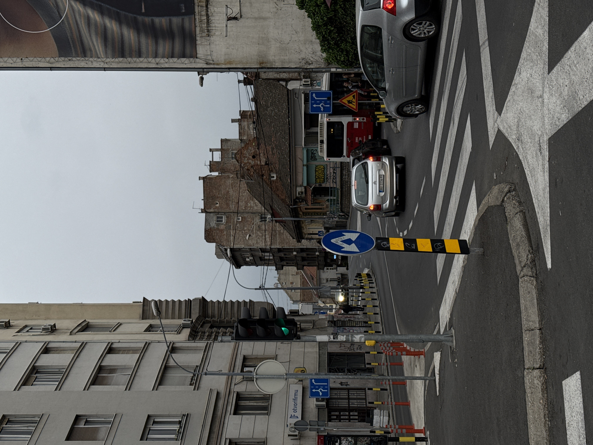 This media file captures an urban street scene in Belgrade, Serbia, under an overcast sky. The image appears rotated 90 degrees clockwise, showing a bustling intersection or road segment.
In the foreground, a silver hatchback car, identified by the Serbian license plate "BG 2236 PV," is visible from the rear, facing away from the viewer. Another silver car is partially visible on the right, facing towards the viewer, positioned near a pedestrian zebra crossing marked with broad white stripes. The road also features various safety barriers and lane dividers, painted in black and yellow stripes. A traffic light, displaying a green signal, indicates permitted traffic flow.
Along the street, a mix of old and more modern buildings lines the thoroughfare. On the left, a multi-story building with multiple windows and air conditioning units stands. Further down the street, older brick buildings with intricate roofing and satellite dishes are visible. A red and white city bus, with "1392" on its rear, is seen between the silver car and the older buildings. A few indistinct figures, likely pedestrians, are visible near the bus.
Several street signs are present, including blue circular mandatory turn signs – one indicating "straight or left turn" and another indicating "mandatory right turn or straight ahead." A yellow triangular warning sign depicting a person shoveling suggests road construction or maintenance nearby. Text visible on a shop front sign reads "F otvorifirmU", possibly relating to business services. The overall impression is that of an active city street on a typical, gray day.