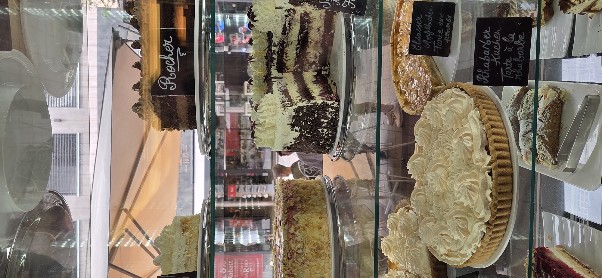The image shows a display case filled with various cakes and pastries. In the foreground, there is a large tart topped with swirls of cream. To its right is an apple tart with a sign that reads "Elässer Apfelkuchen. Tarte aux pommes". To the left of the cream tart is a slice of red cake with visible layers. Behind these, on a higher shelf, is a chocolate cake with cream frosting, labeled "Rocher". Next to it, a partially sliced cake with white and dark chocolate layers and cherries on top is visible. Further back and to the left on the top shelf is a slice of white cake. The display case appears to be in a bakery or cafe. Through the glass, people can be seen inside the establishment and walking on a street. The overall impression is that of a sweet treat selection offered to customers. The mention of Kehl, Germany in the prompt suggests the location of this establishment.