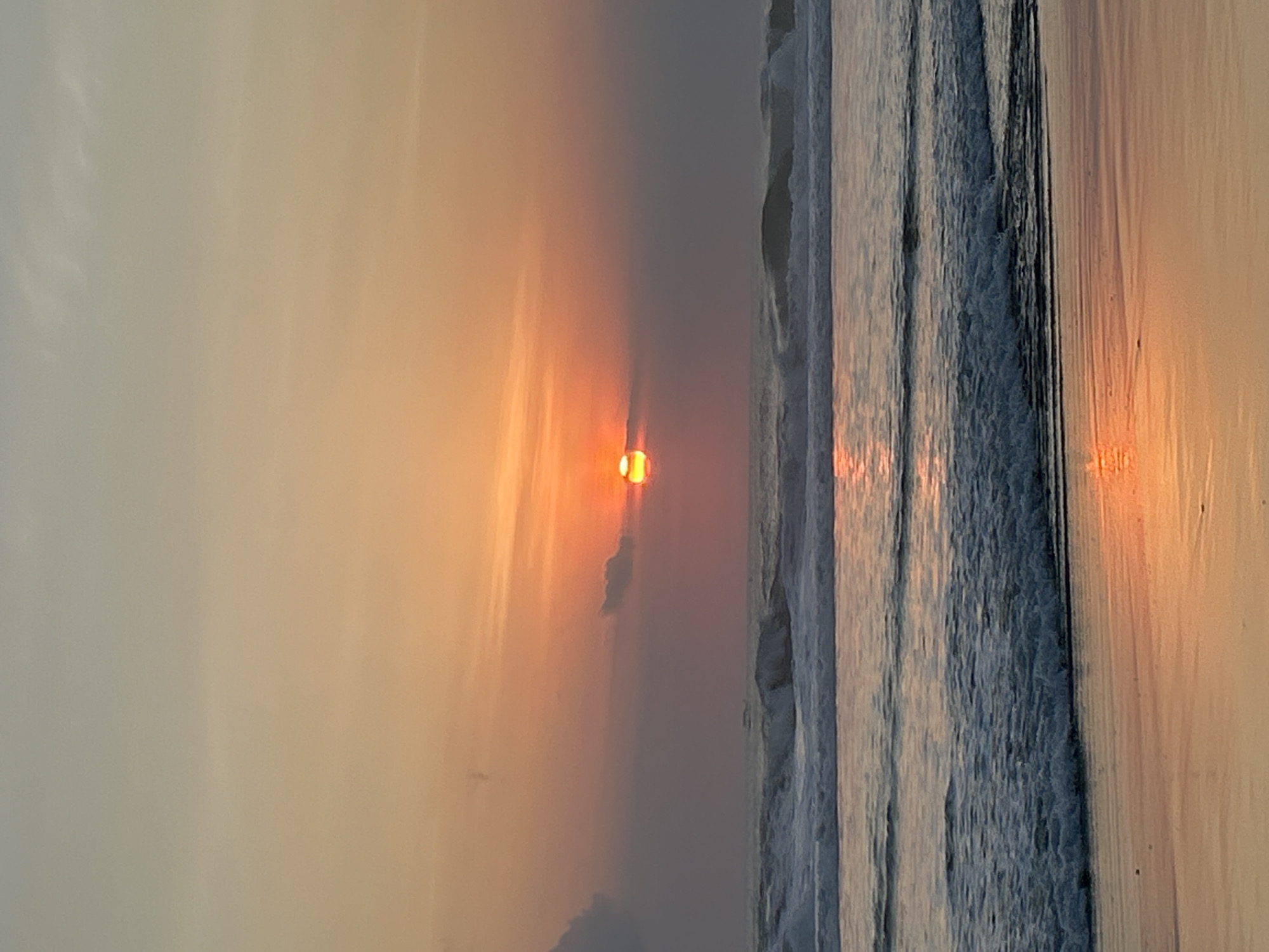 The image captures a serene sunset over the ocean in North Kuta, Indonesia. The sky is a gradient of soft orange and yellow hues, with the sun, a vibrant orb of red and orange, partially obscured by a wispy cloud. The ocean is calm, with gentle waves rolling towards the shore. The wet sand in the foreground reflects the warm colors of the sky and the setting sun. There are no people or specific structures visible in the frame, emphasizing the natural beauty of the coastal landscape. The overall atmosphere is peaceful and tranquil.