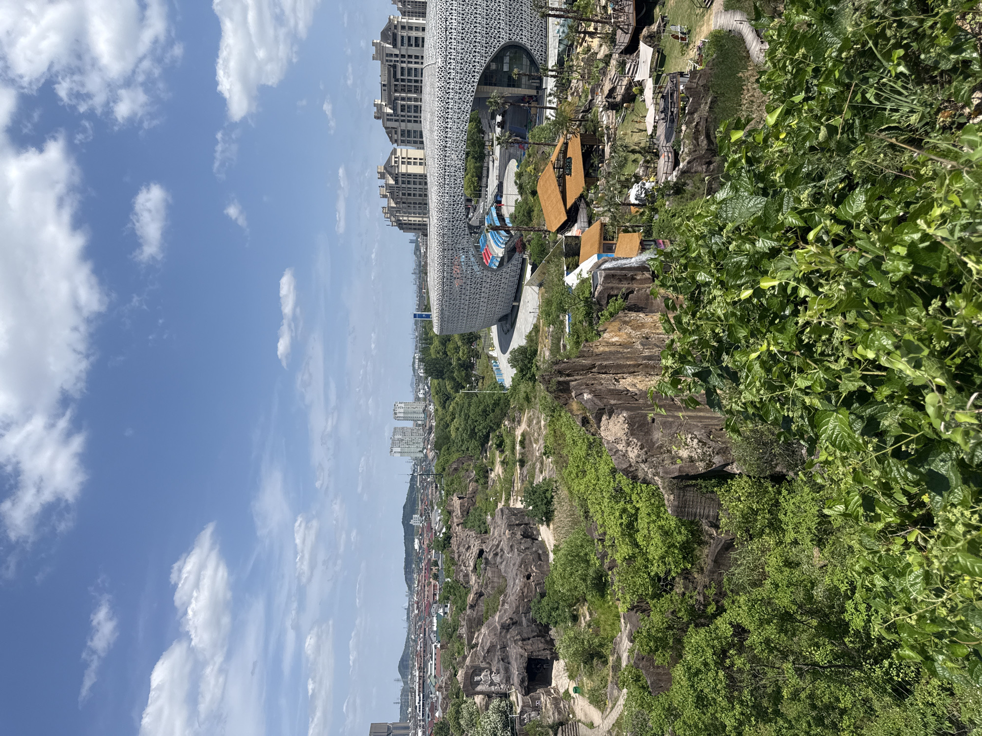 The image presents an overview of a mixed urban and natural environment. In the foreground, there is dense greenery with trees and foliage obscuring the view of the city below. The terrain is rocky and uneven. In the mid-ground, the city is visible with a mix of residential buildings, commercial structures, and green spaces. Notable is a large, modern building with a white latticed facade. This building has a distinctive rounded shape and appears to be a cultural or entertainment venue. Further back, the cityscape extends toward the horizon with taller buildings and structures. The sky is bright blue with scattered white clouds. The location appears to be the city of Keqiao District, China, evidenced by the style of architecture and landscaping.