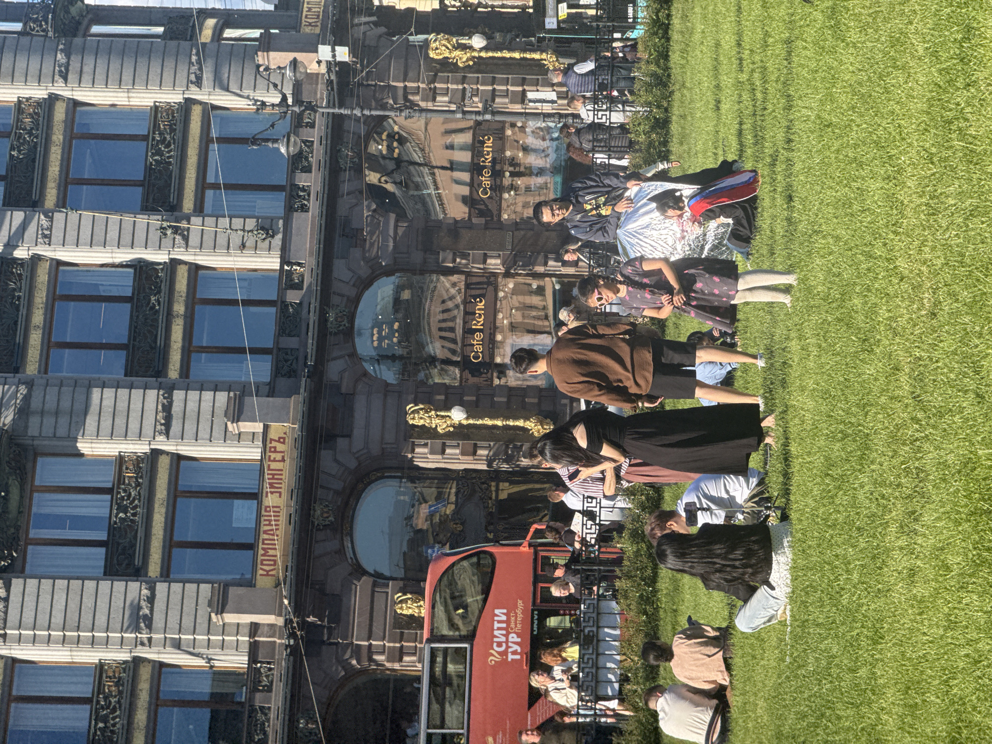 This media file captures a sunny day in Saint Petersburg, Russia, with a group of people gathered on a vibrant green lawn in front of a grand, historic building. The building features ornate architectural details, including decorative elements above arched windows and a long banner with Cyrillic text. The text on the banner reads "КОМПАНИЯ ЗИНГЕРЪ" which translates to "Singer Company." Another prominent sign visible on the building's facade says "Cafe René."
In the foreground, a bright red double-decker tour bus with "СИТИ ТУР" (City Tour) written on it is partially visible, suggesting a tourist activity. Several people are interacting with each other on the grass. Some are sitting, while others appear to be standing or walking. The overall atmosphere seems relaxed and casual, indicative of a pleasant day outdoors. The sunlight suggests it is daytime, likely mid-morning or afternoon, and the clear skies imply fair weather. The people's clothing varies, from casual t-shirts to dresses and jackets, reflecting a range of personal styles. Some individuals are looking at their phones, while others are engaged in conversation. The composition of the image, with the architectural grandeur of the building contrasting with the natural expanse of the lawn, creates a visually appealing scene that blends urban and recreational elements.