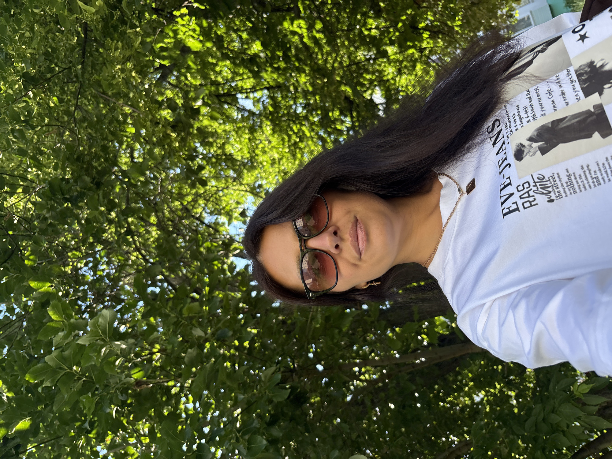 A woman with long, dark hair and sunglasses is pictured from the chest up, outdoors. She is wearing a white t-shirt with a graphic print and a gold necklace. Behind her, a dense canopy of green leaves from a tree filters the sunlight. The image appears to have been taken in a park or garden setting.